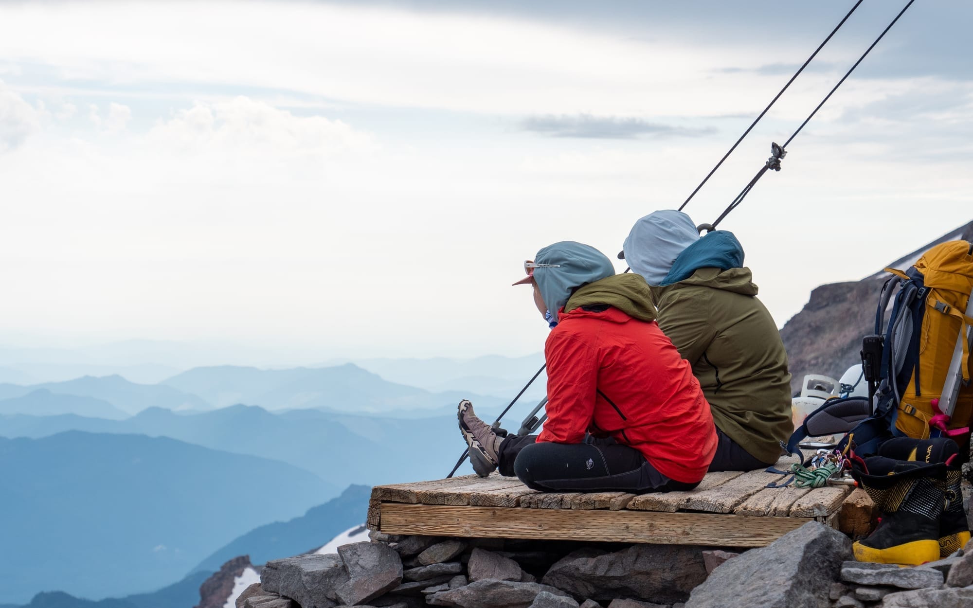 Two hikers sitting on a wooden platform overlooking misty mountain layers under a cloudy sky.