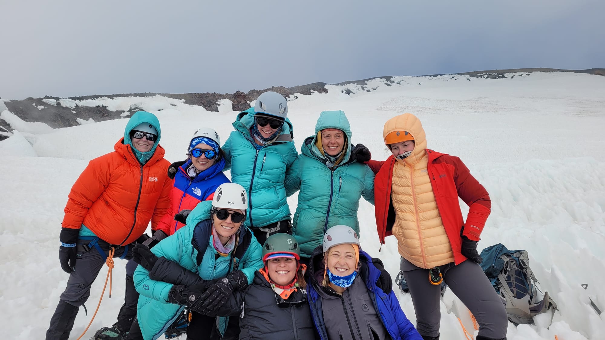 Group of eight women climbers in winter gear posing happy on a snowy mountain glacier.