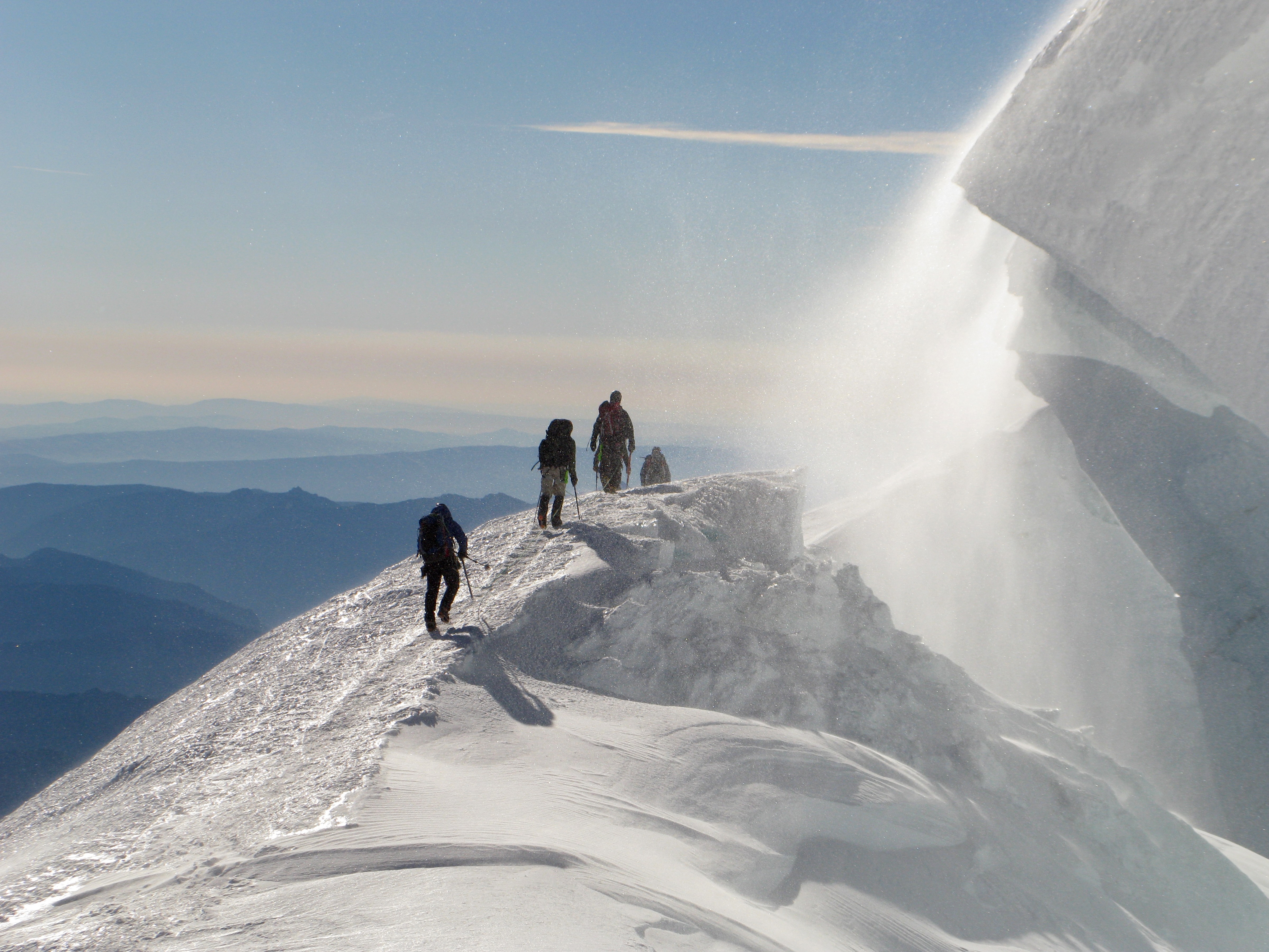 Four climbers hiking along a snow ridge near a blowing snow cornice on a mountain peak.