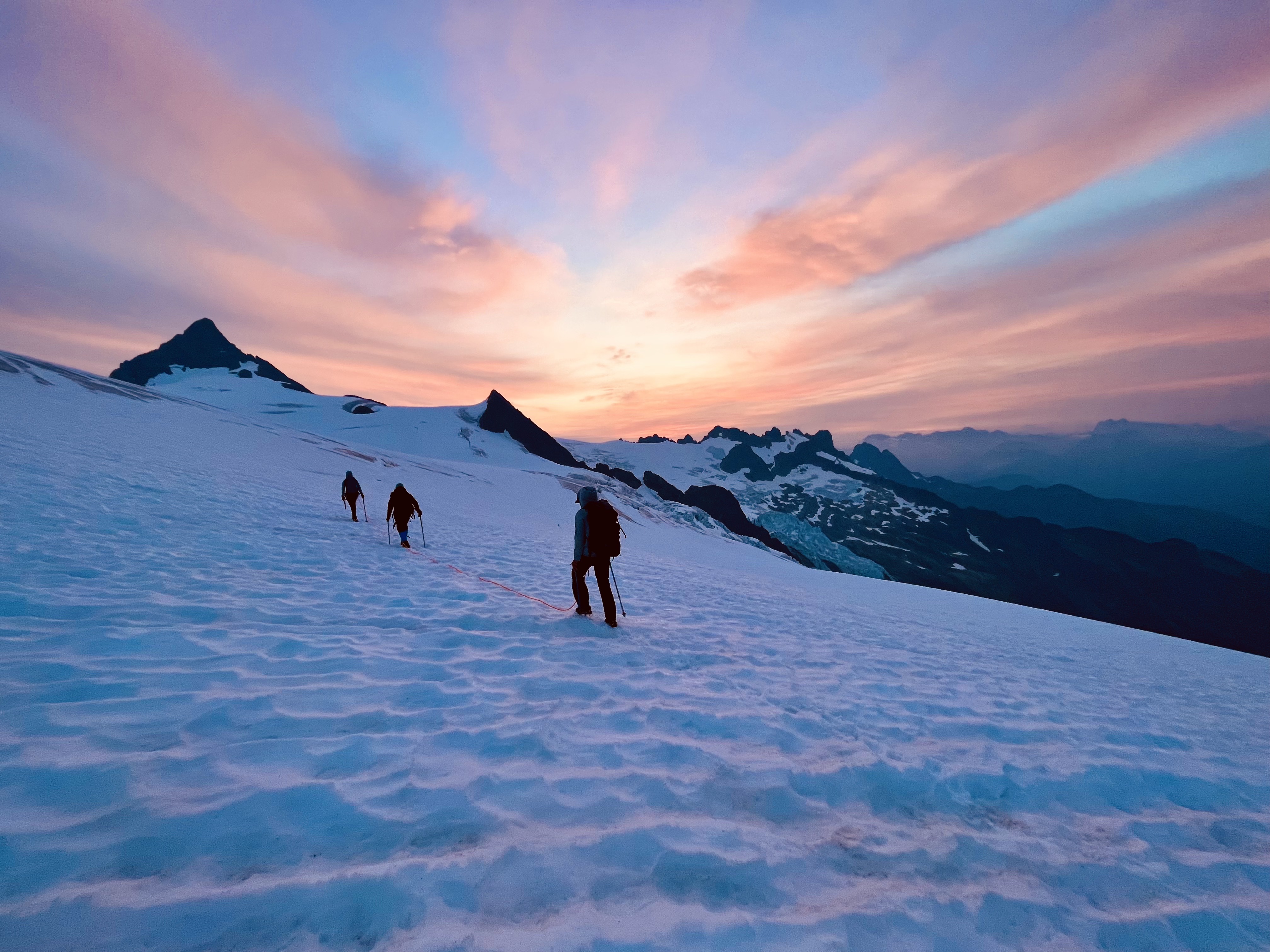 Three climbers roped together ascending snowy mountain slope at vibrant sunrise or sunset.