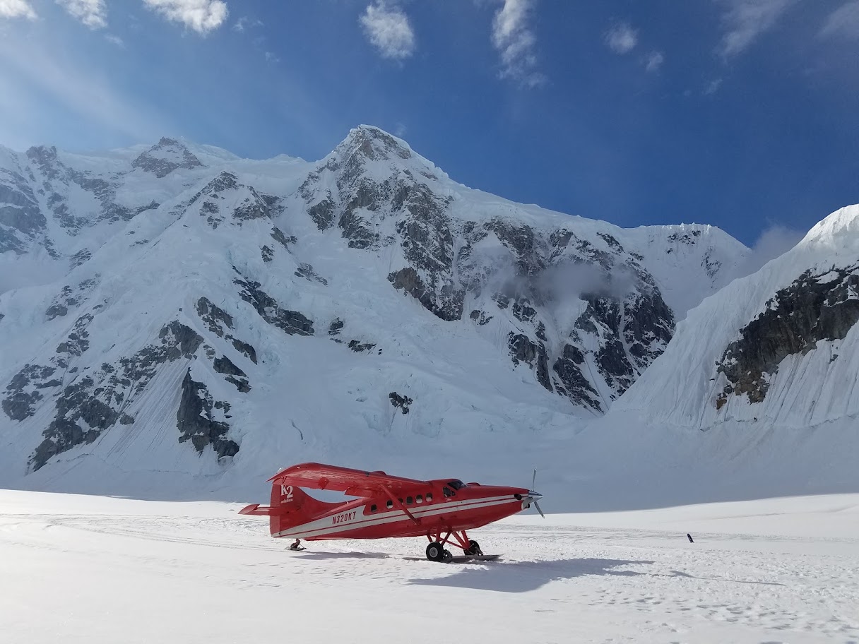 Red K2 airplane parked on snowfield in front of tall snowy mountain under blue sky.