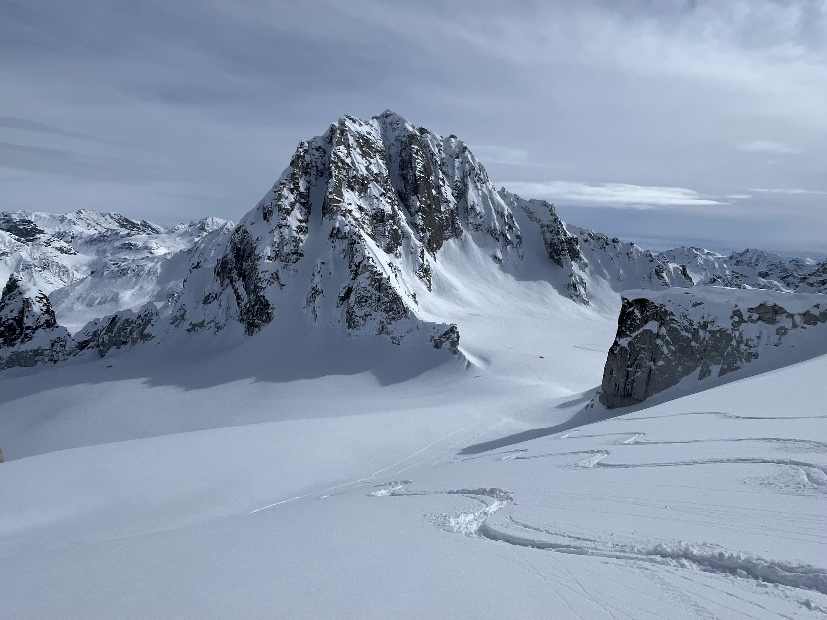 Snow-covered rugged mountain with ski tracks in the foreground under a cloudy sky.