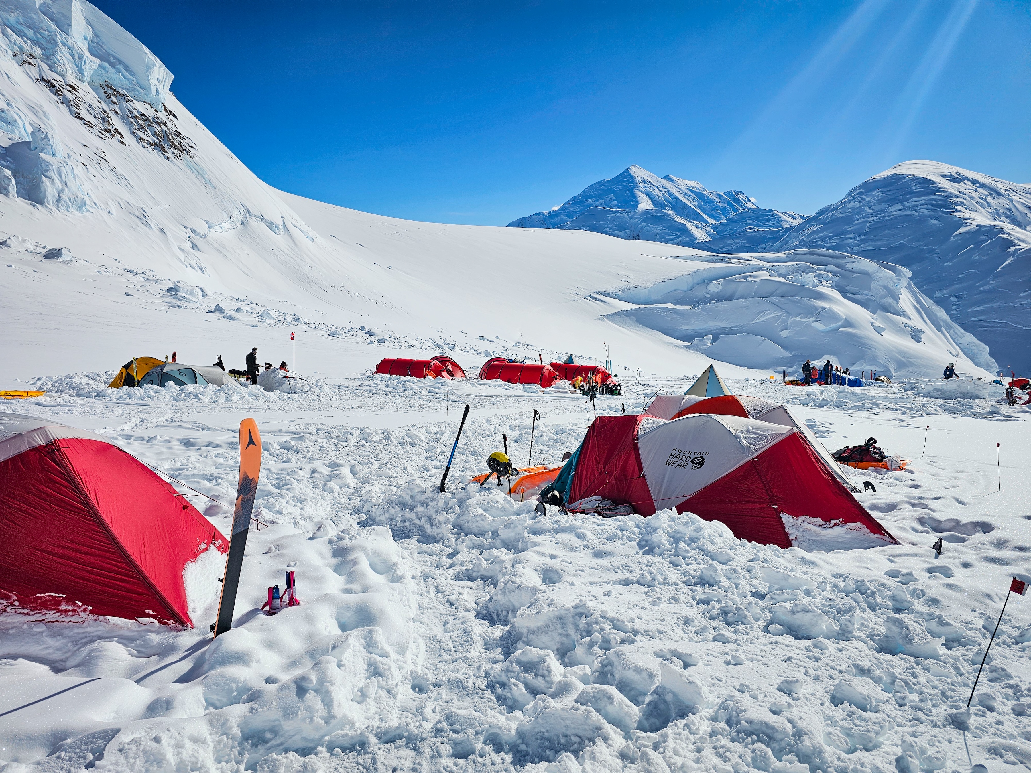 Snow-covered base camp with red tents and ski gear under bright blue sky and mountains.