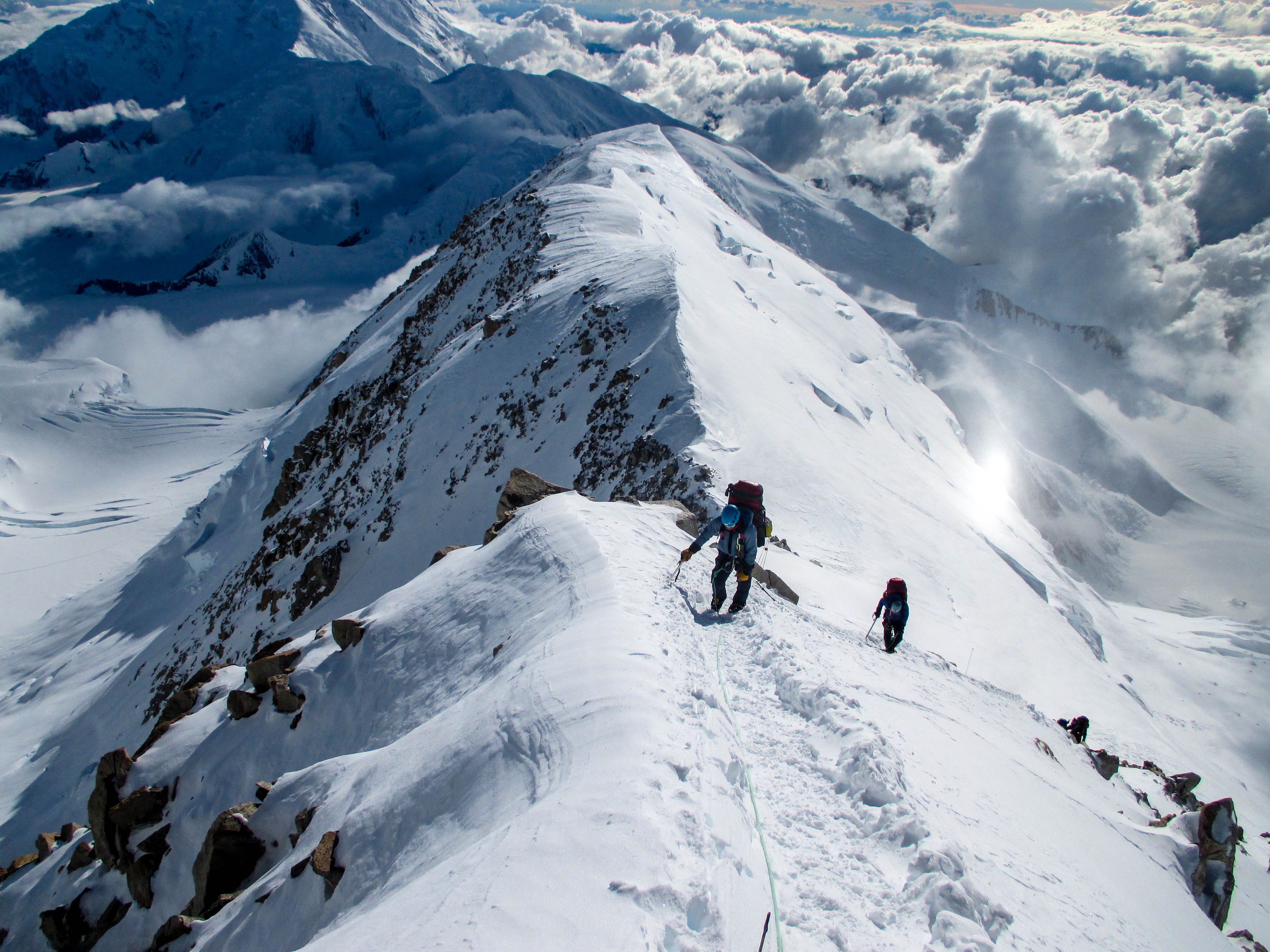 Three climbers ascending a snowy mountain ridge high above clouds and rugged peaks.