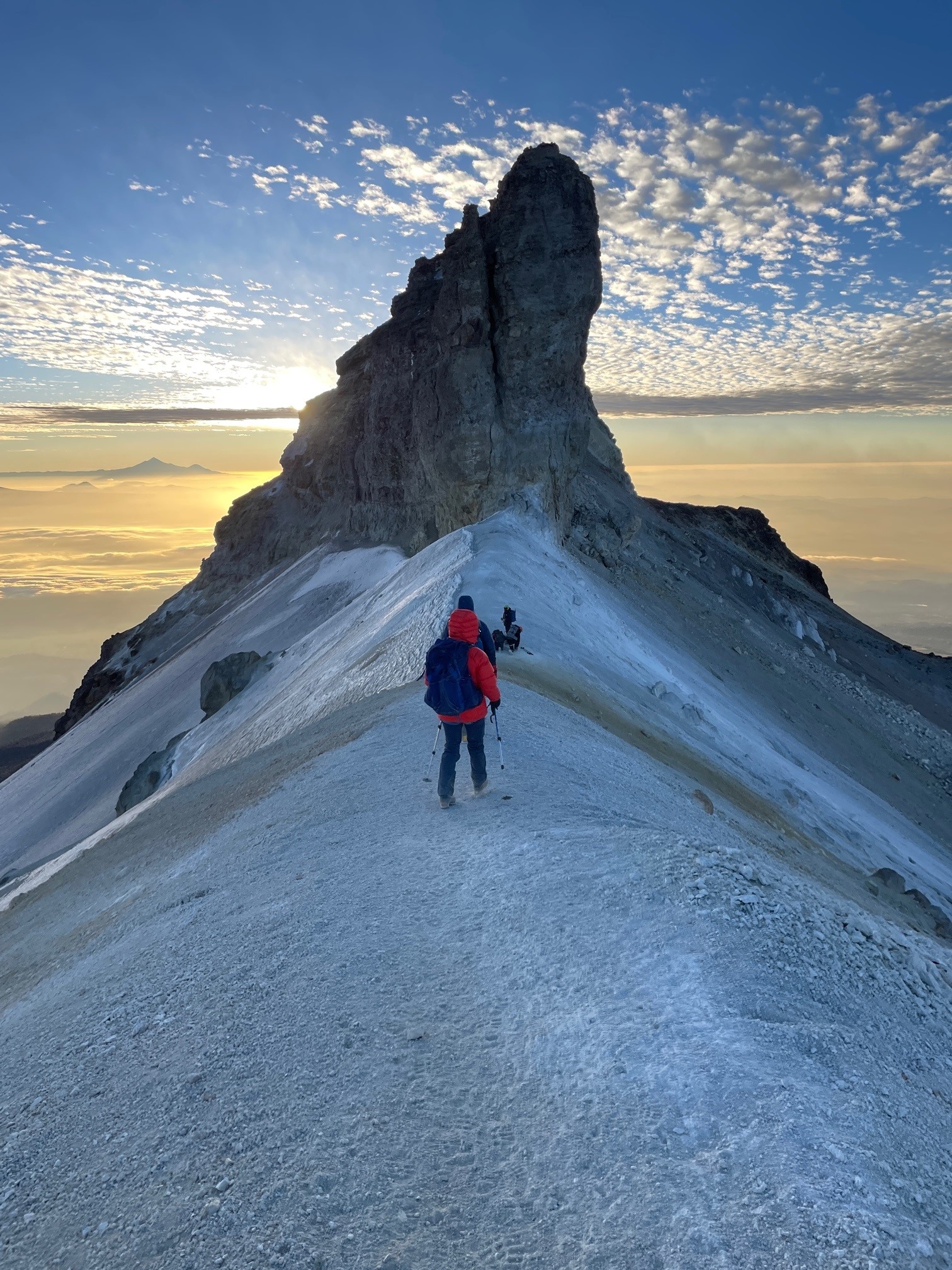 Hikers climbing a snowy ridge toward a tall rocky peak at sunrise with a cloudy sky.