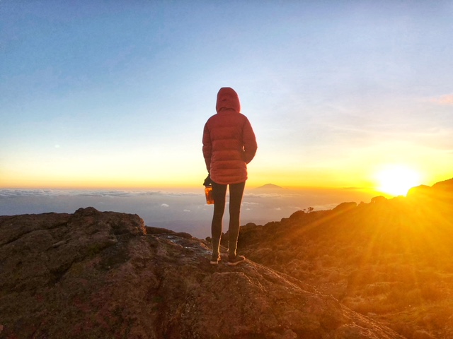 Person in red jacket standing on rocky hill watching sunrise over distant mountains and clouds.