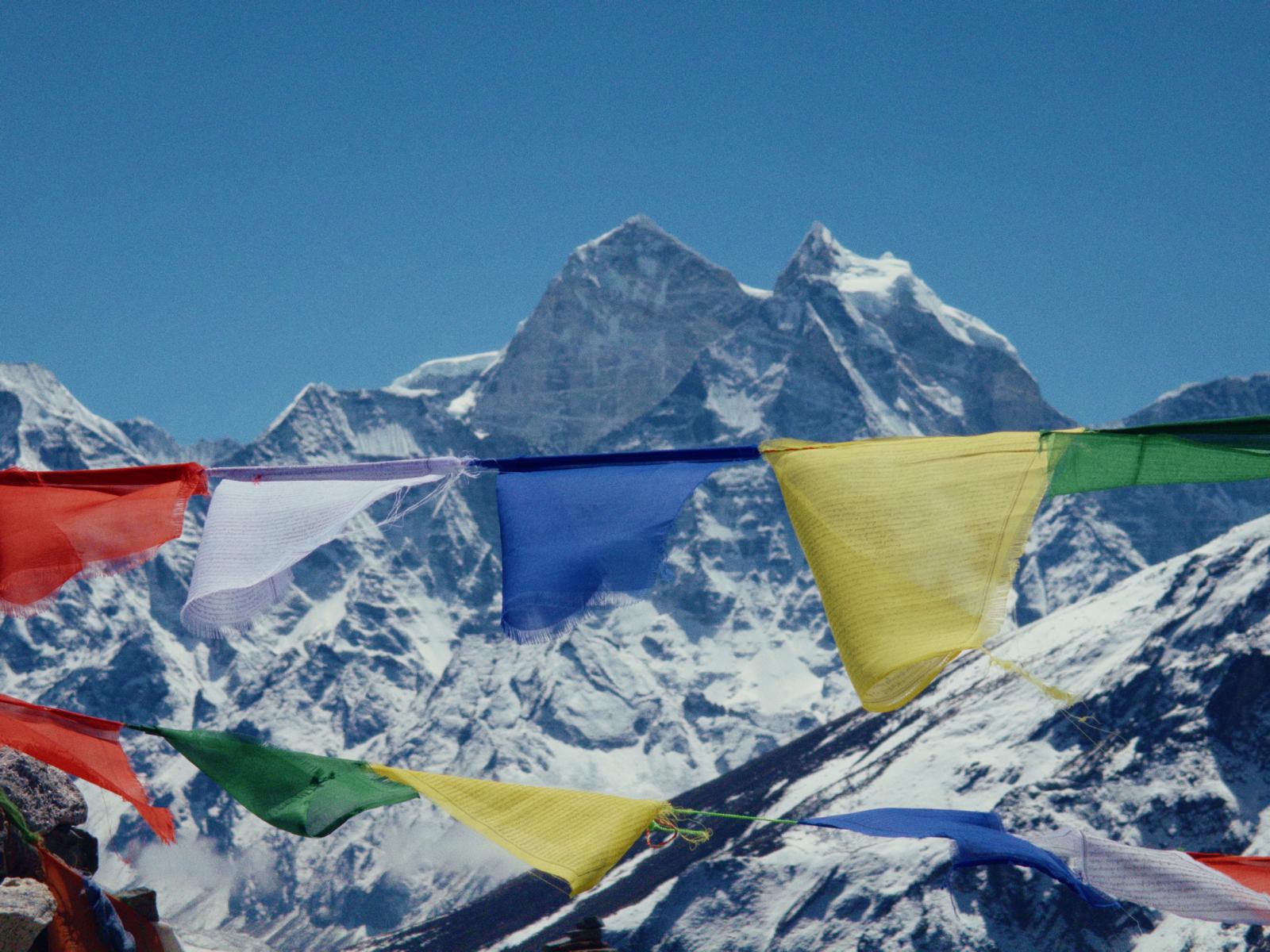 Colorful prayer flags fluttering in front of snow-covered Himalayan mountain peaks under a clear blue sky.