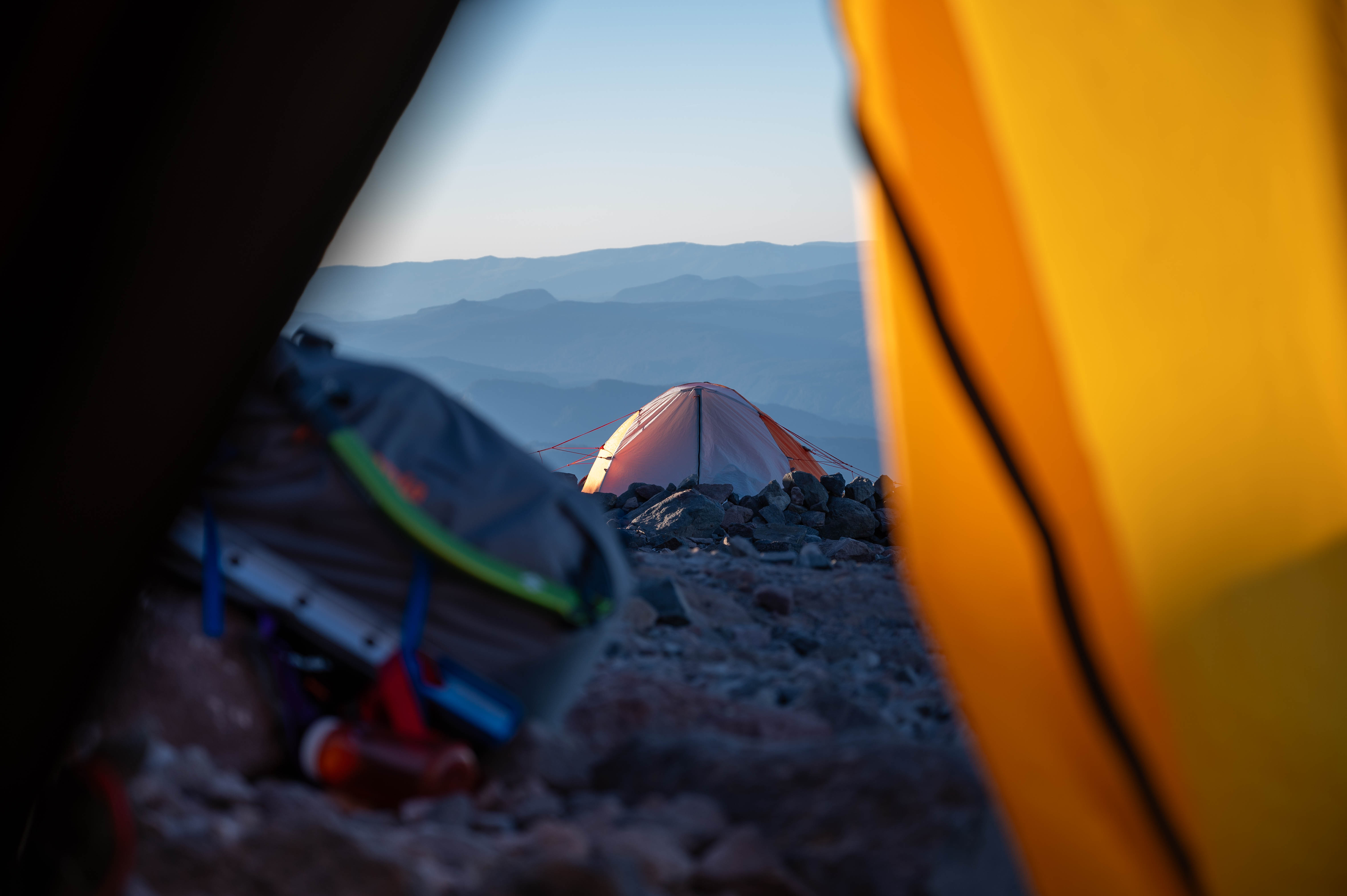 Orange tent pitched on rocky ground framed by black and yellow tent flaps with blue mountains in background.