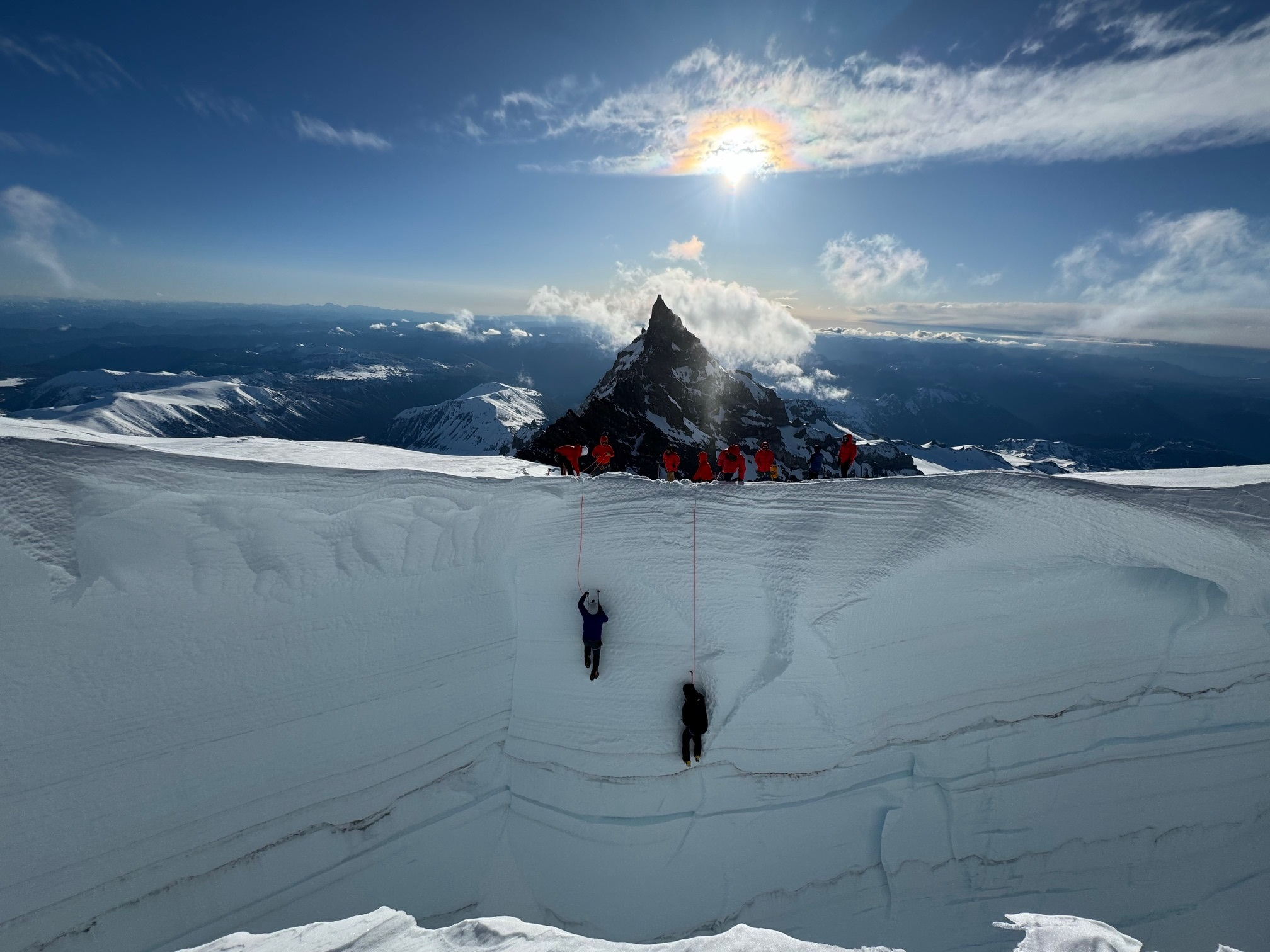 Climbers in red jackets assist two people climbing a steep snowy crevasse on a mountain peak.