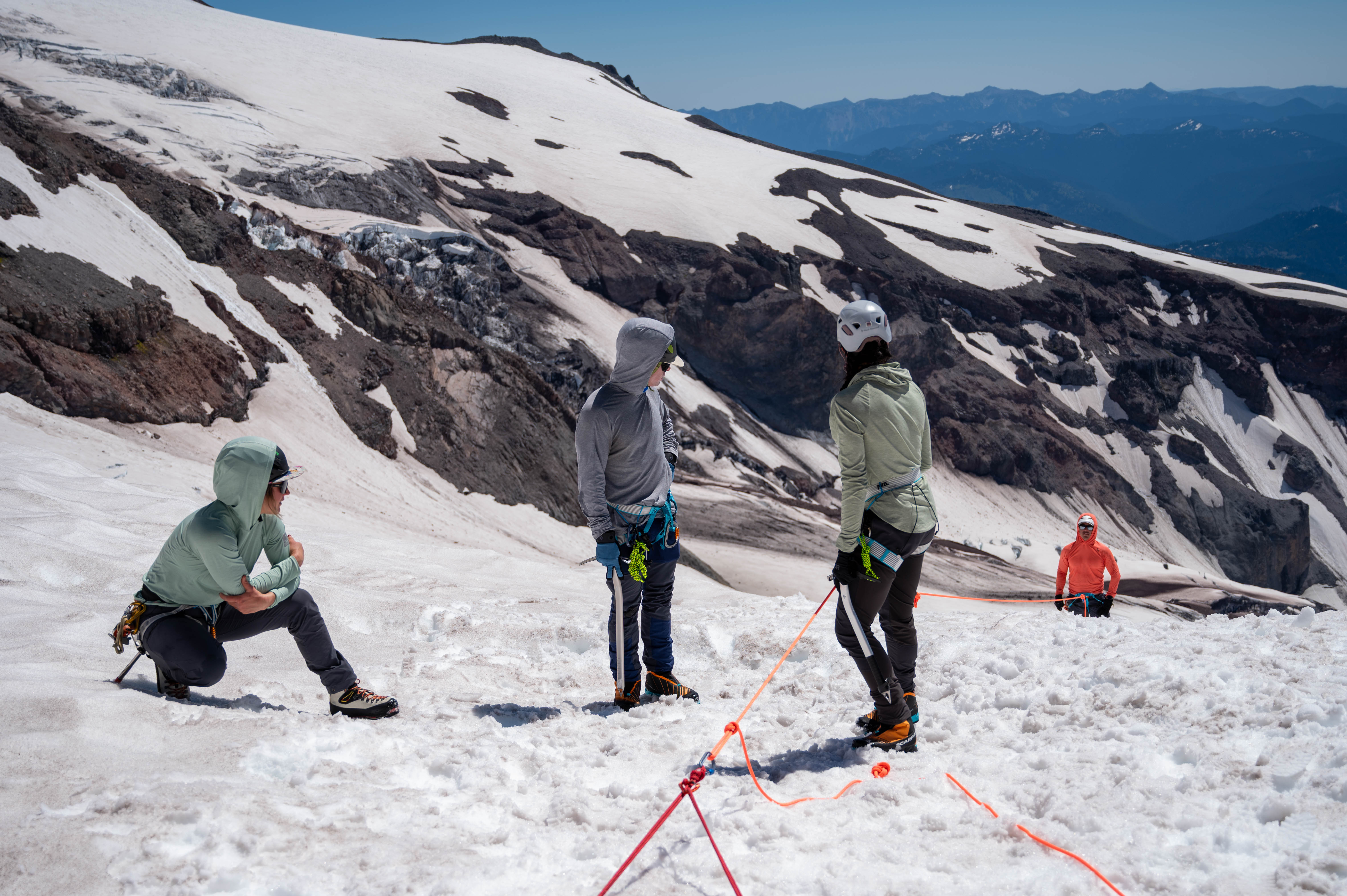 Four climbers with ice axes on snowy mountain slope practicing rope safety techniques.
