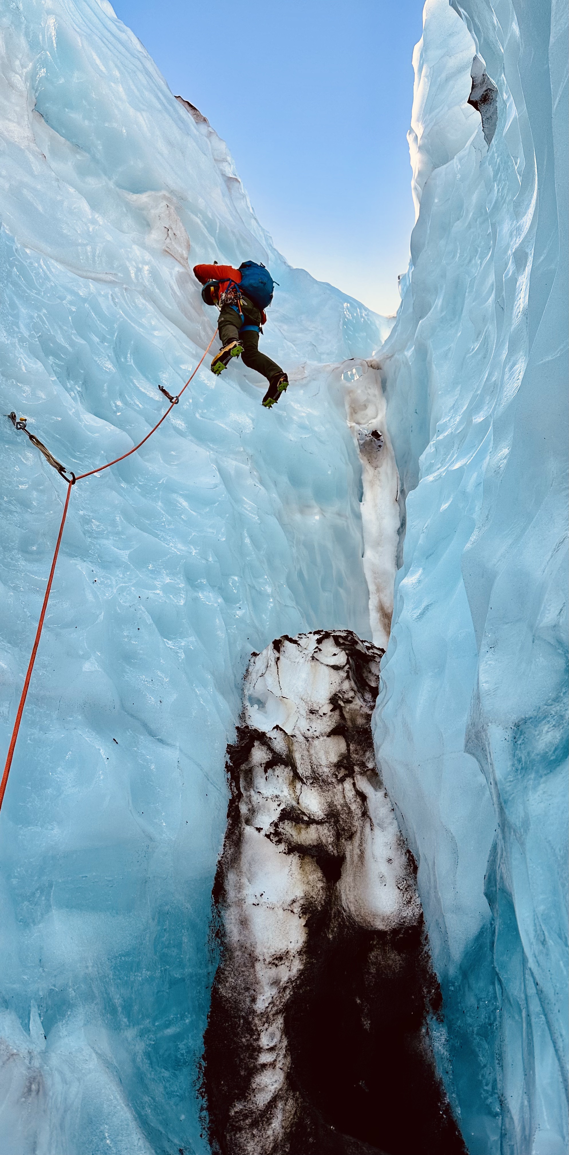 Climber in red jacket ascending an icy blue glacier crevasse with safety rope and climbing gear.