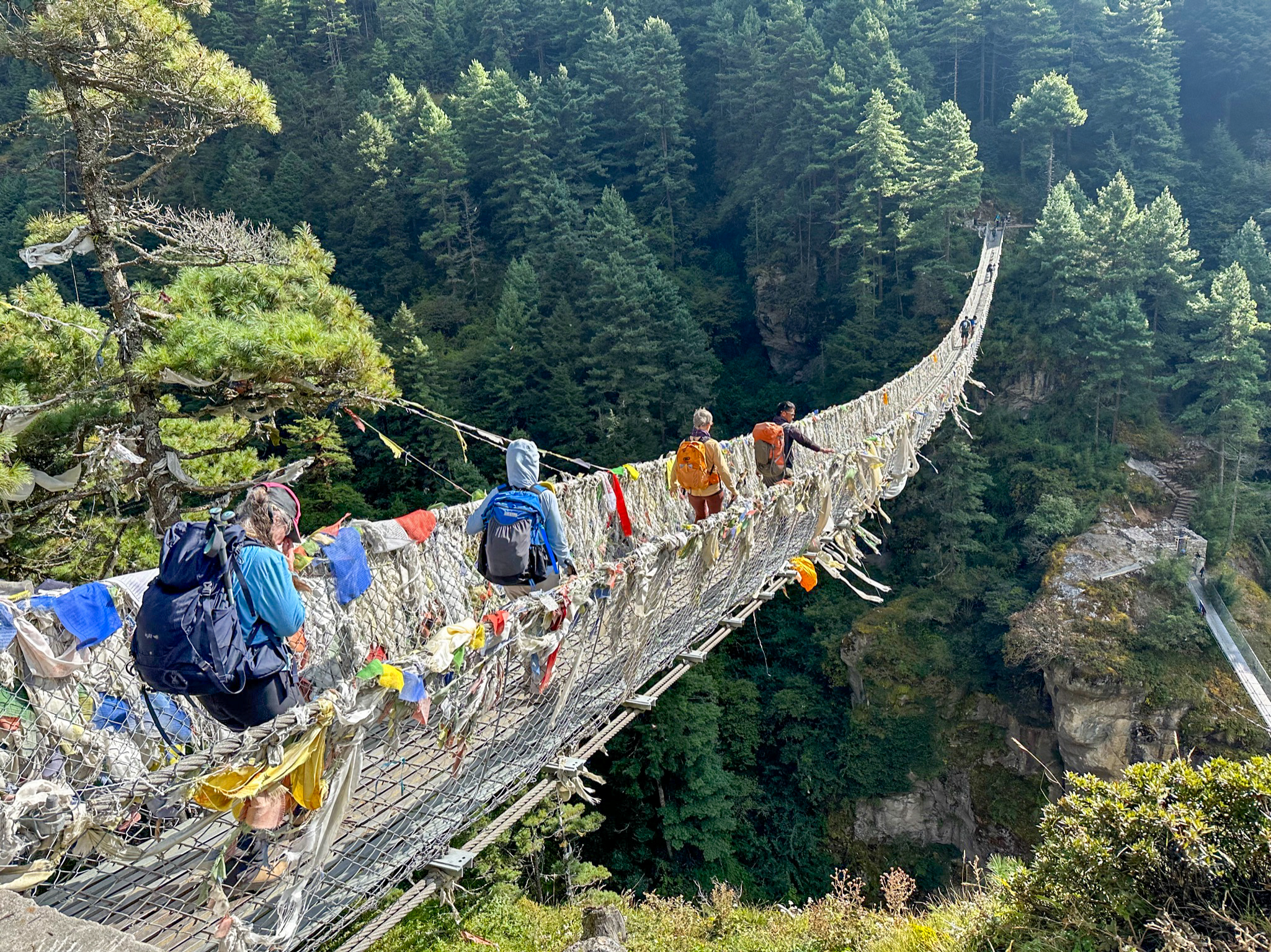 Hikers with backpacks crossing a suspension bridge covered with prayer flags over a forested gorge.