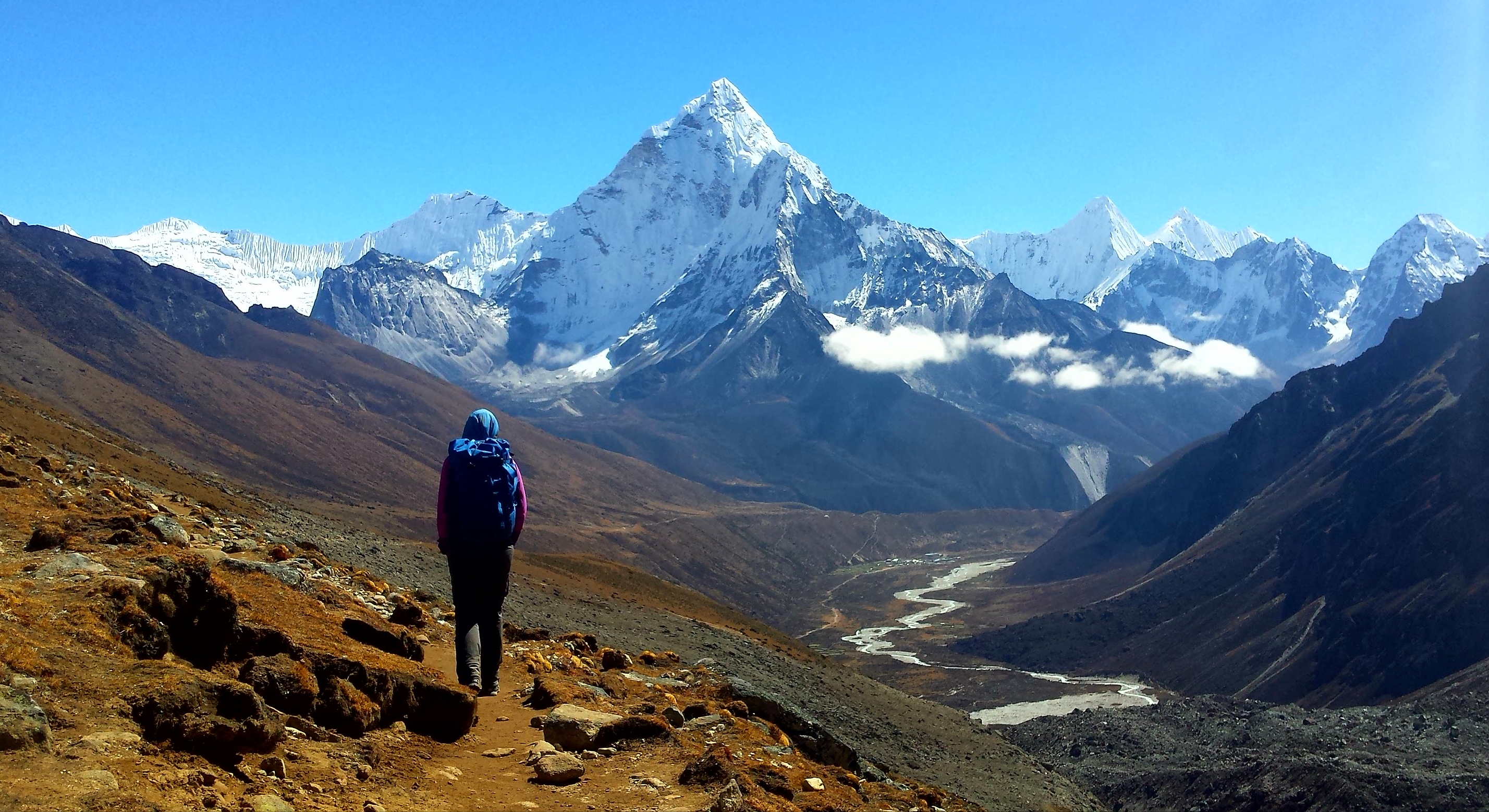 Person hiking on mountain trail with snow-covered peaks and winding river in a clear blue sky.
