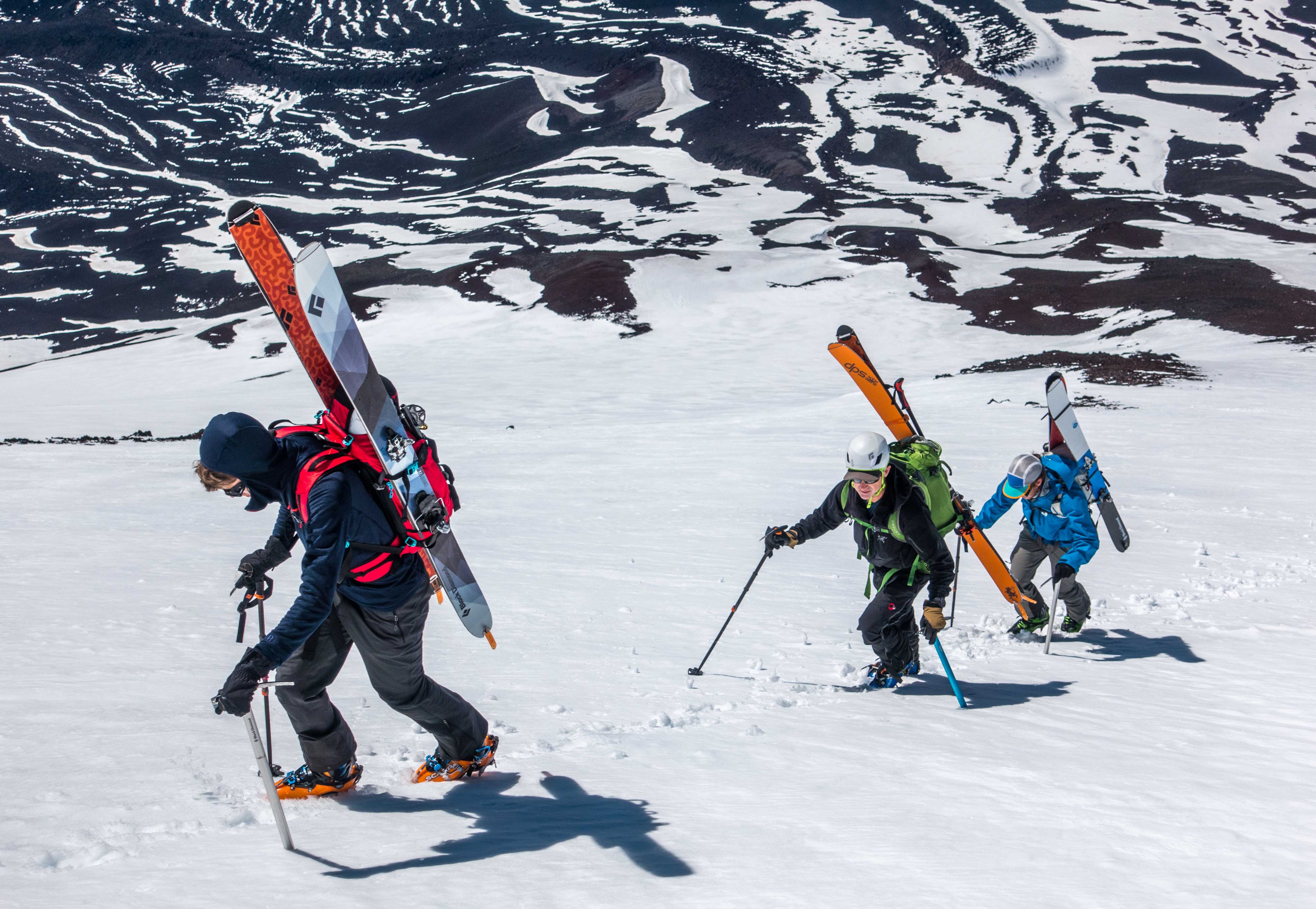 Three climbers hiking up a snowy mountain carrying skis and wearing backpacks and helmets.