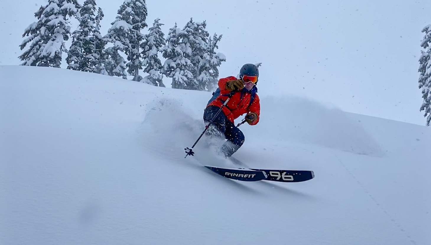 Skier in orange jacket and helmet making a turn in fresh powder snow with snowy trees in background.