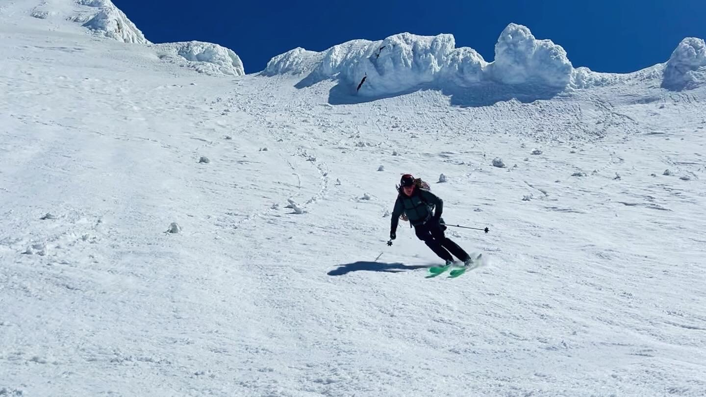 Skier in dark clothing with green skis carving turns down a snowy mountain slope under clear blue sky.