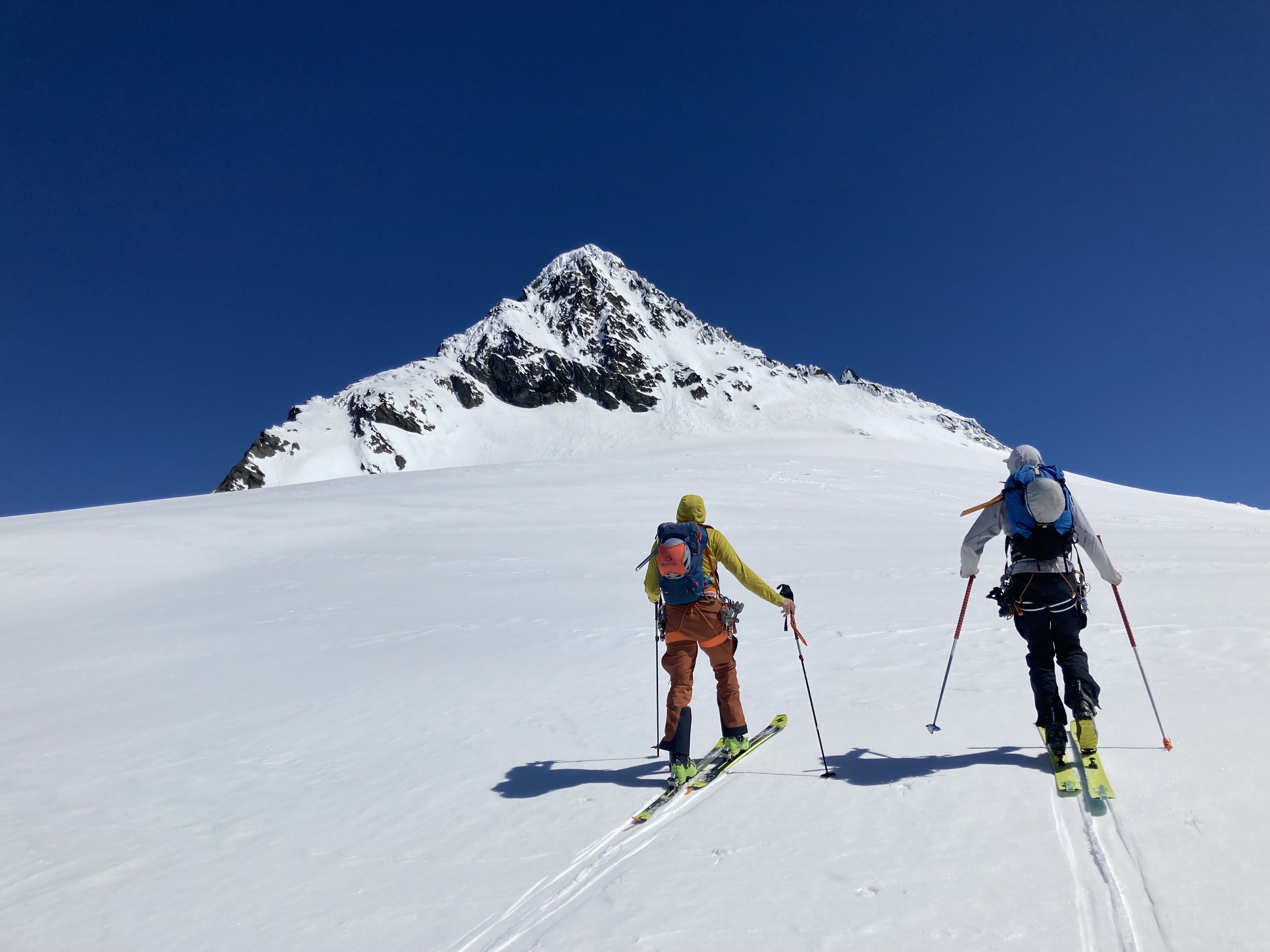 Two skiers ascending snowy mountain slope with a steep, rocky peak under clear blue sky.