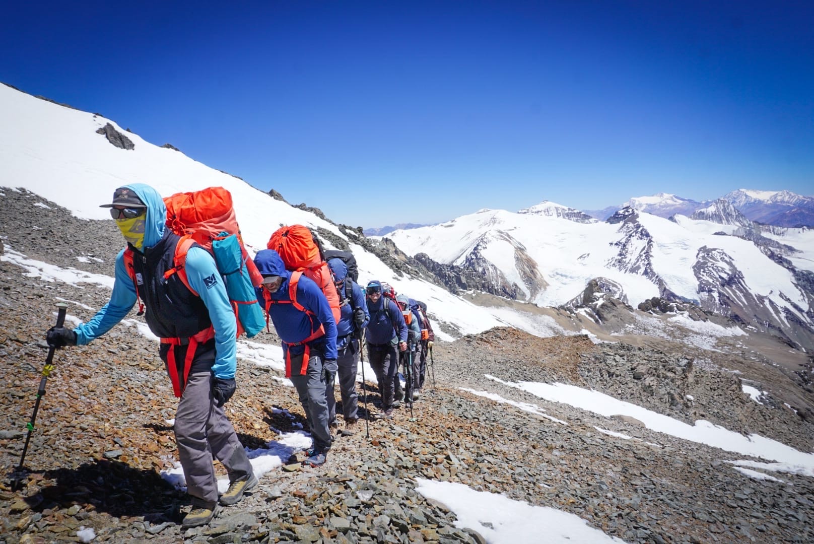 Hikers in blue jackets with orange backpacks trekking in a rocky snowy mountain landscape.