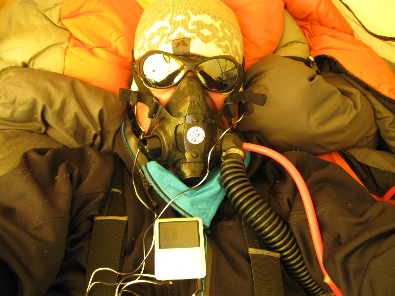 Person wearing a gas mask, sunglasses, and warm clothing with earphones and a music player.