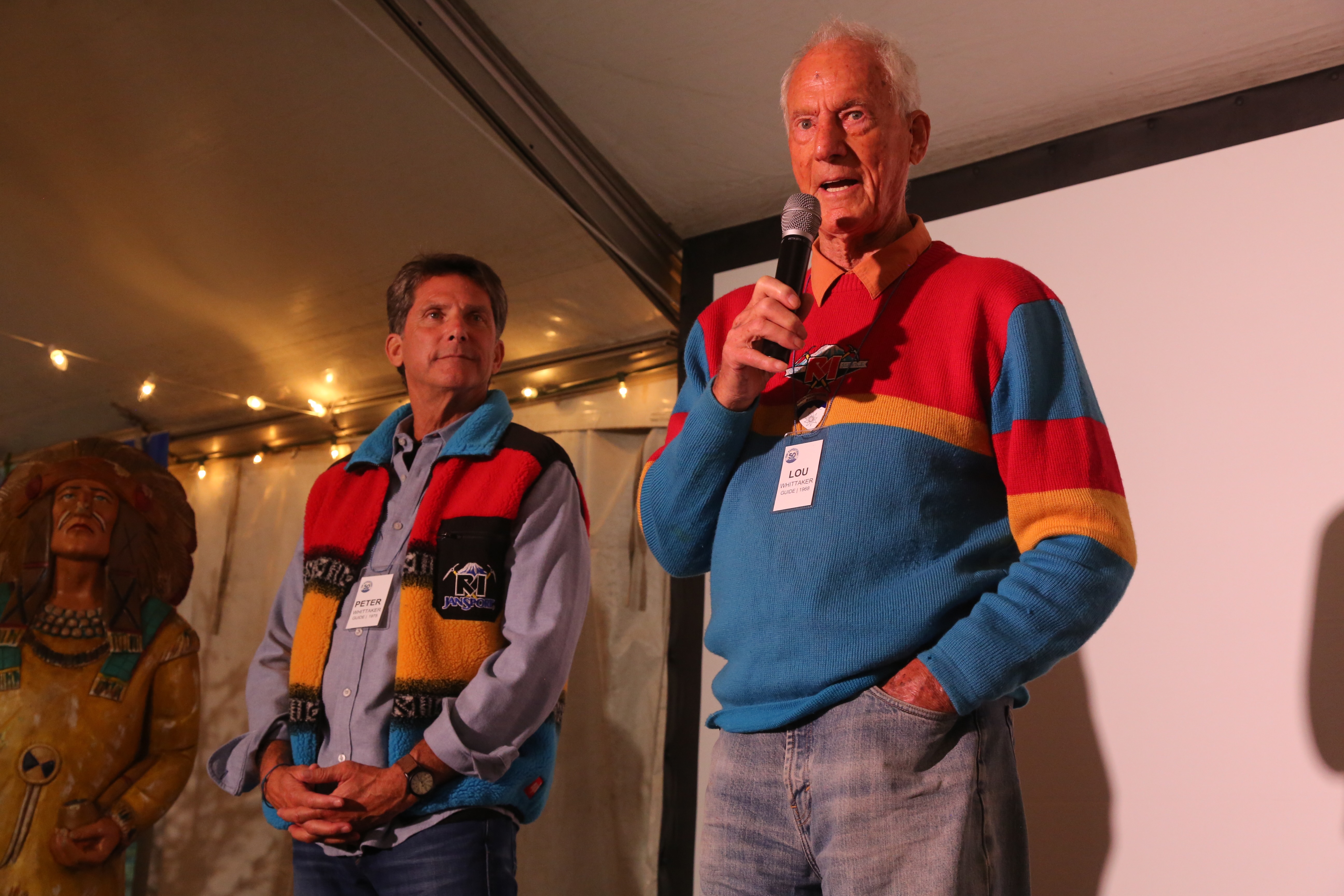 Two men wearing colorful sweaters and name tags speak inside a tent with string lights.