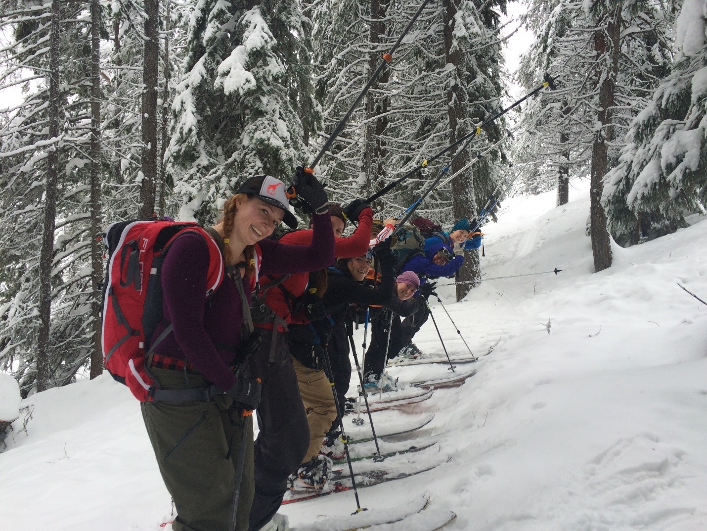 Group of six skiers posing with poles raised on snowy trail surrounded by snow-covered trees.