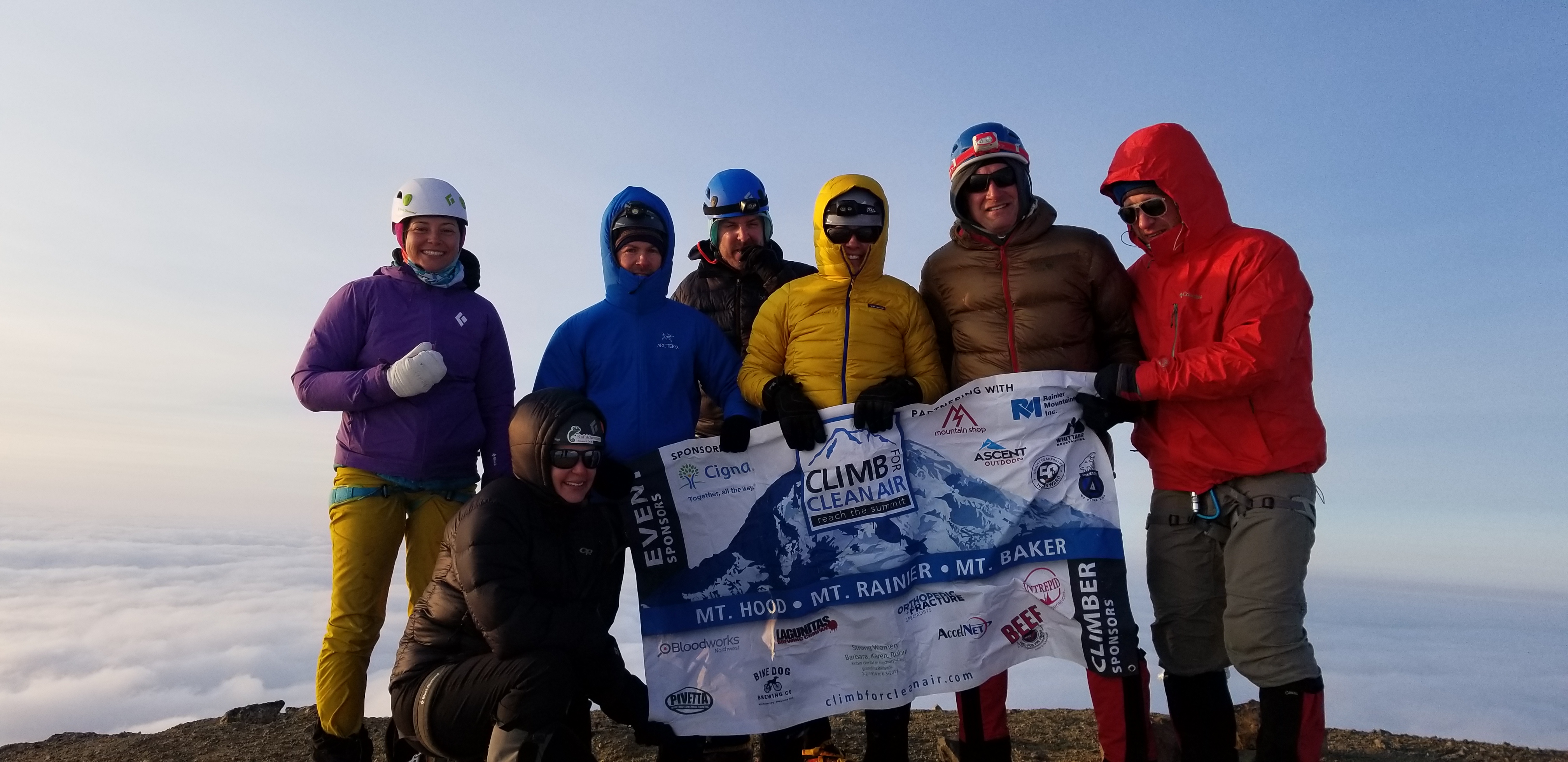 Seven climbers in outdoor gear holding a Climb for Clean Air banner atop a mountain above clouds.