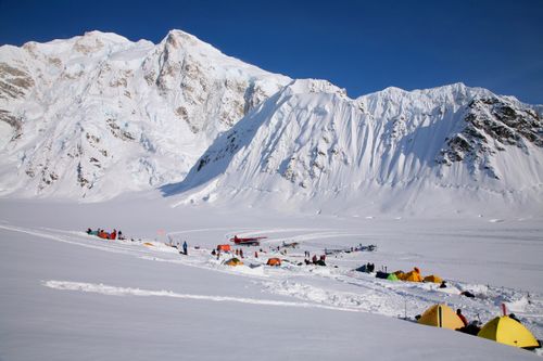 Snow-covered camp with tents and small planes at the base of large snowy mountains under a clear blue sky.