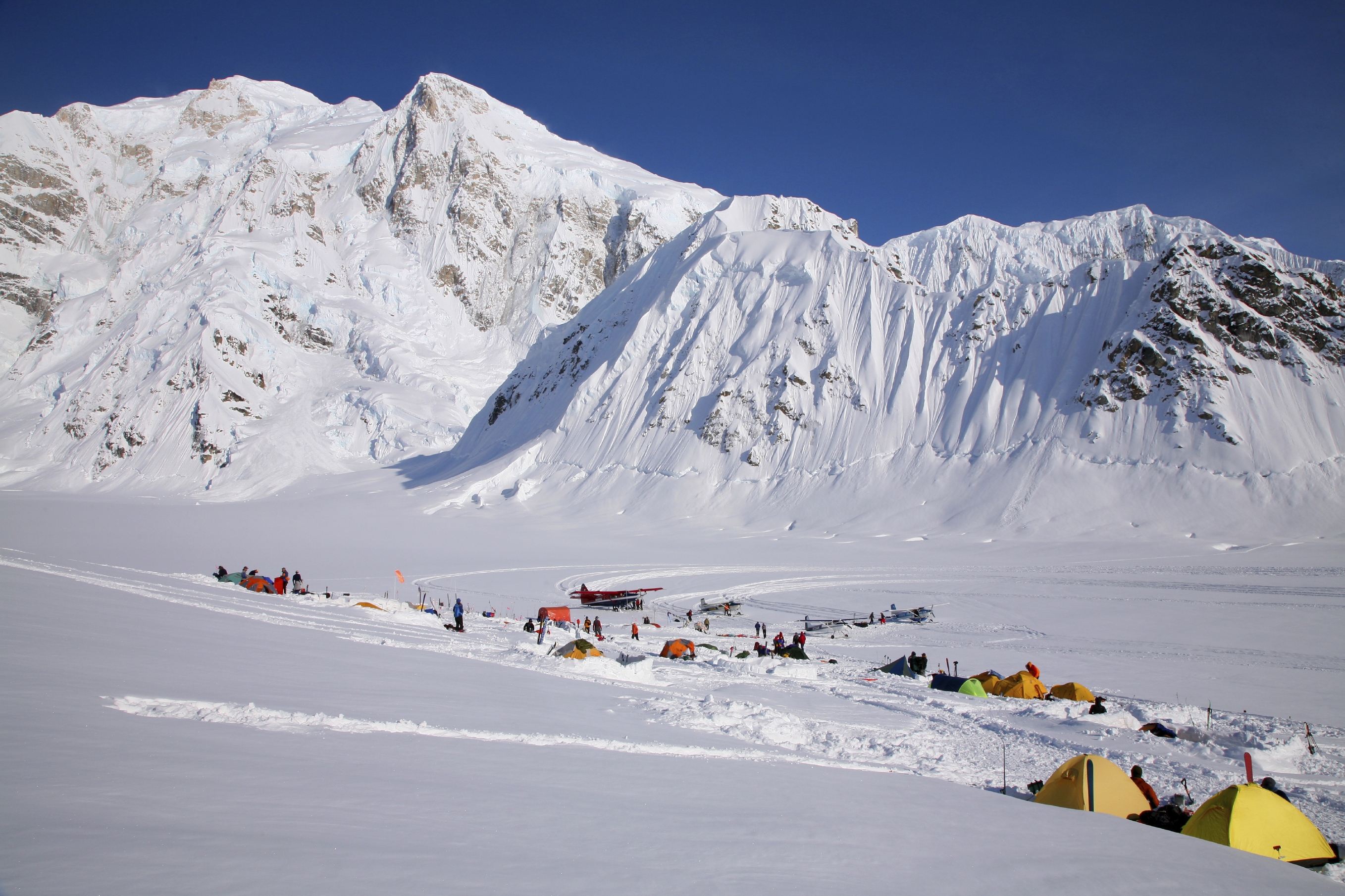 Snow-covered camp with tents and small planes at the base of large snowy mountains under a clear blue sky.
