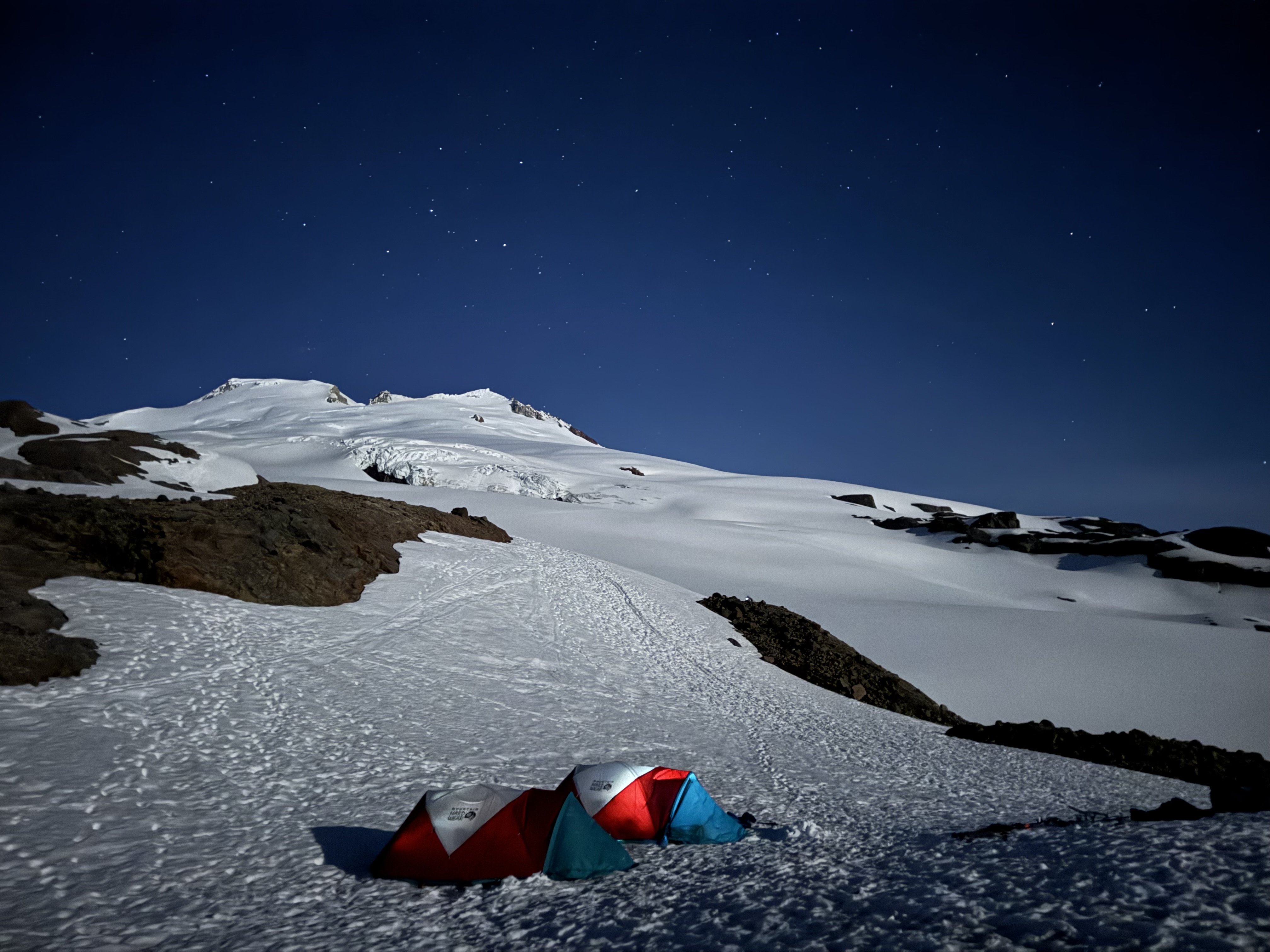 Three colorful tents pitched on snow at night under a starry sky with snowy mountain slopes.