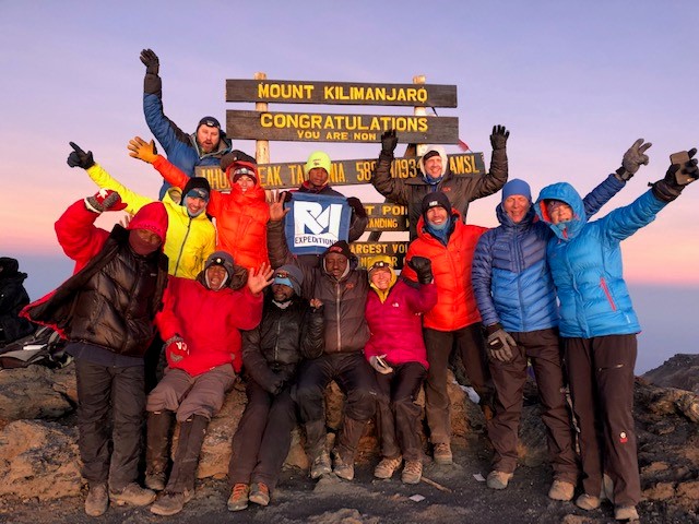 Group of climbers celebrating at Mount Kilimanjaro summit sign during sunrise.