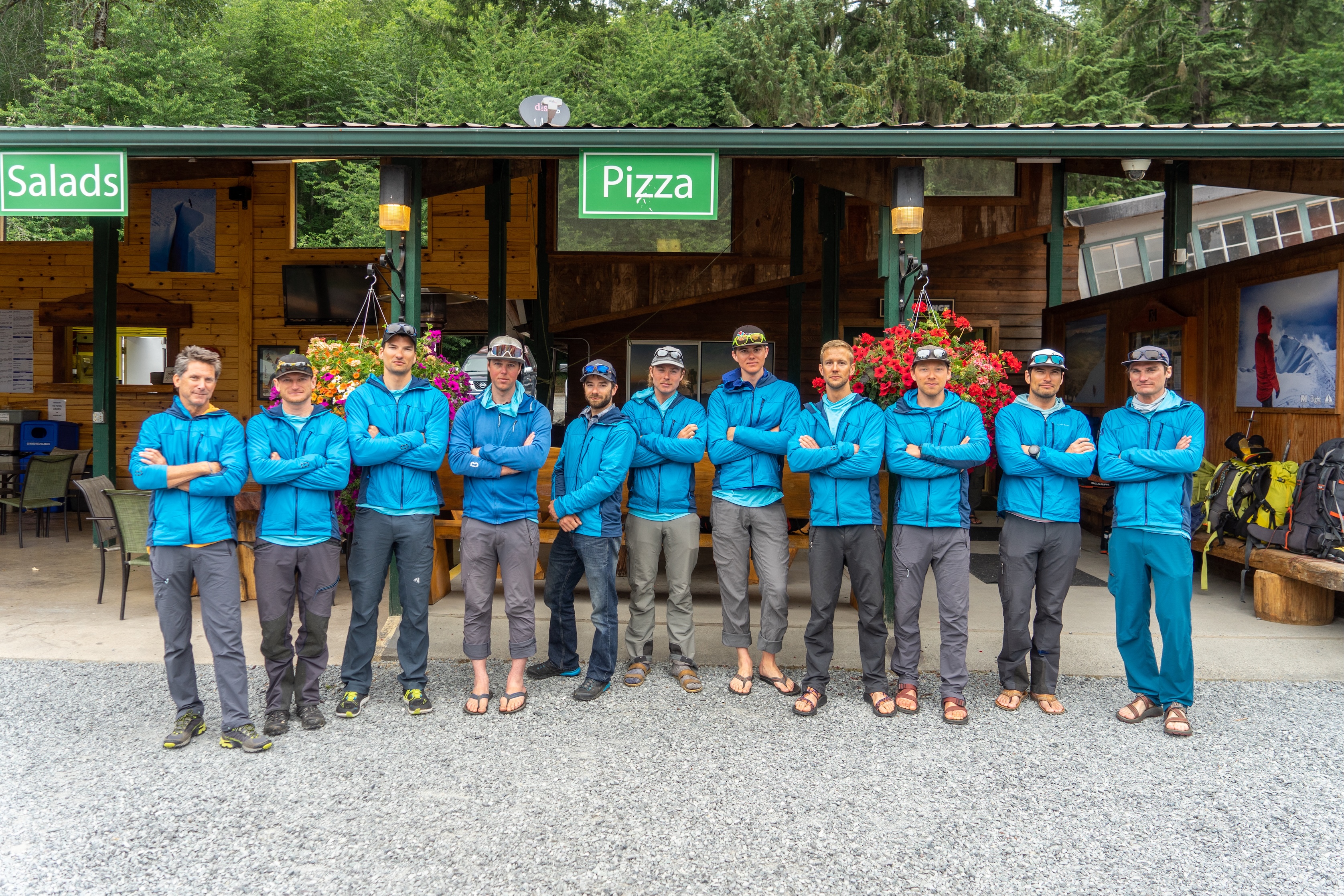 Eleven men in blue jackets stand with arms crossed in front of a wooden building with signs for salads and pizza.