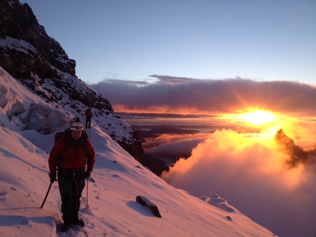 Two climbers in red jackets ascending a snowy mountain slope at sunrise above clouds.