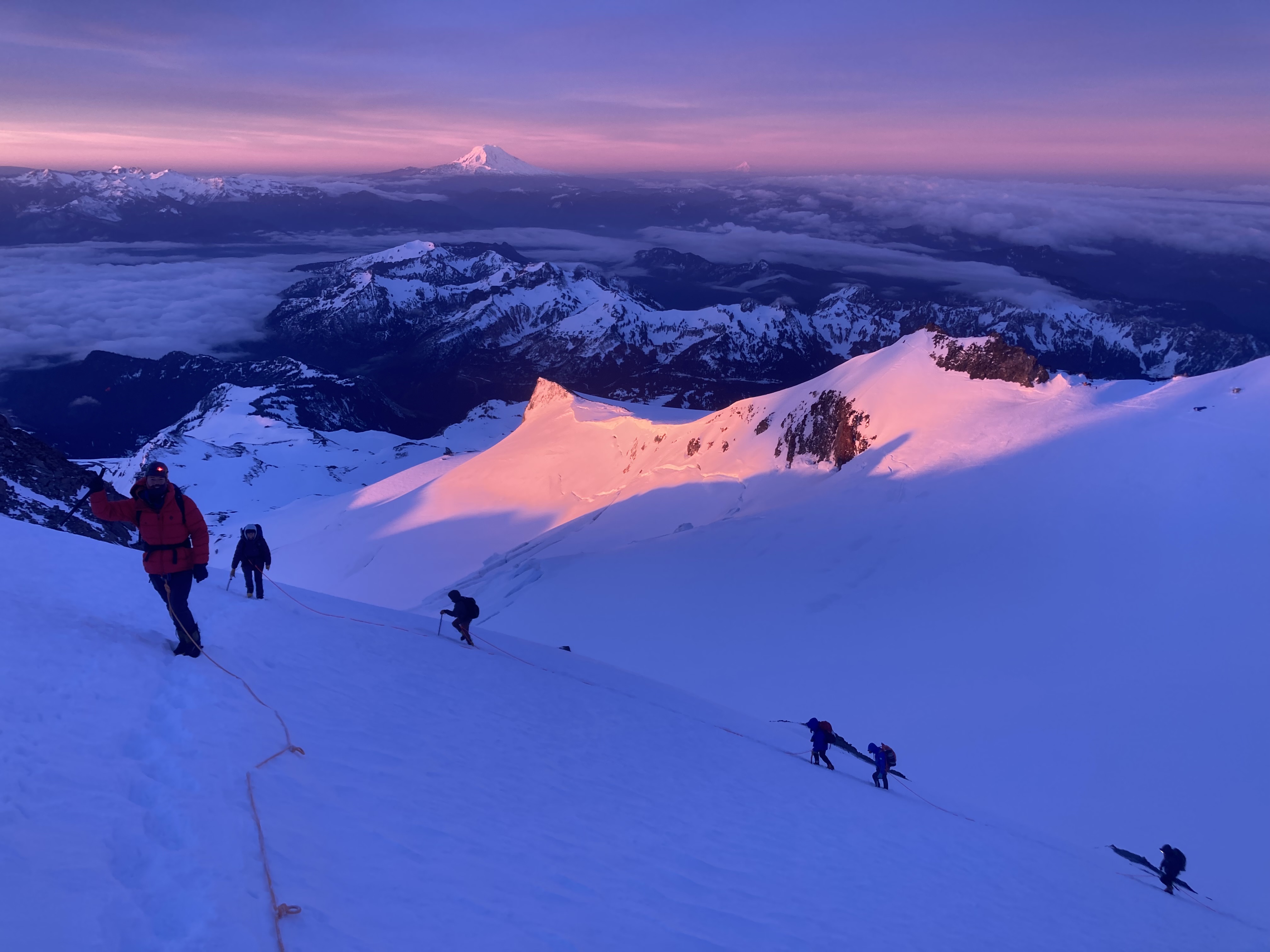 Climbers roped together ascending snowy mountain slope at sunrise with distant peaks and purple sky.