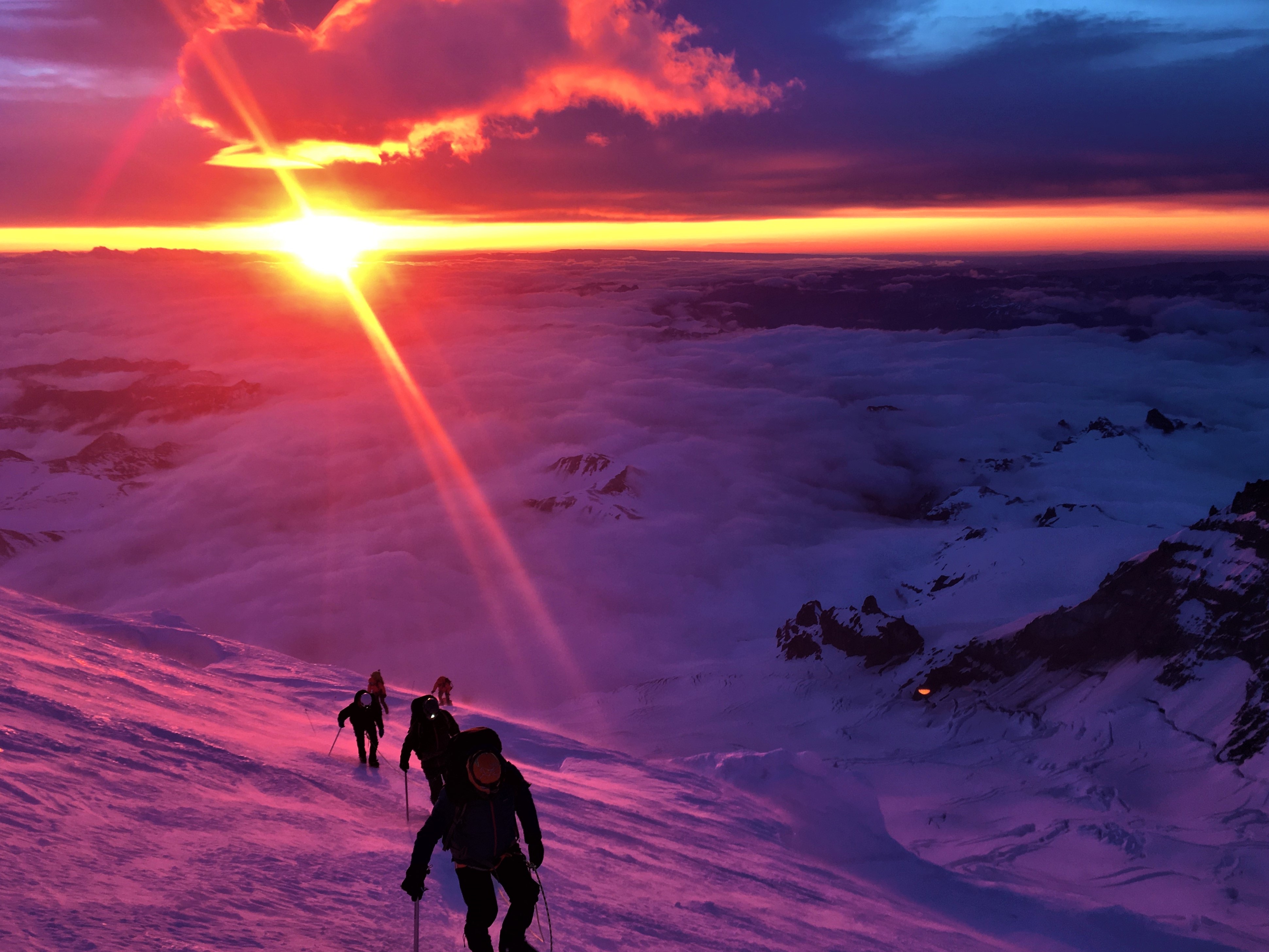 Climbers ascending a snowy mountain slope at sunrise with clouds and mountains below.