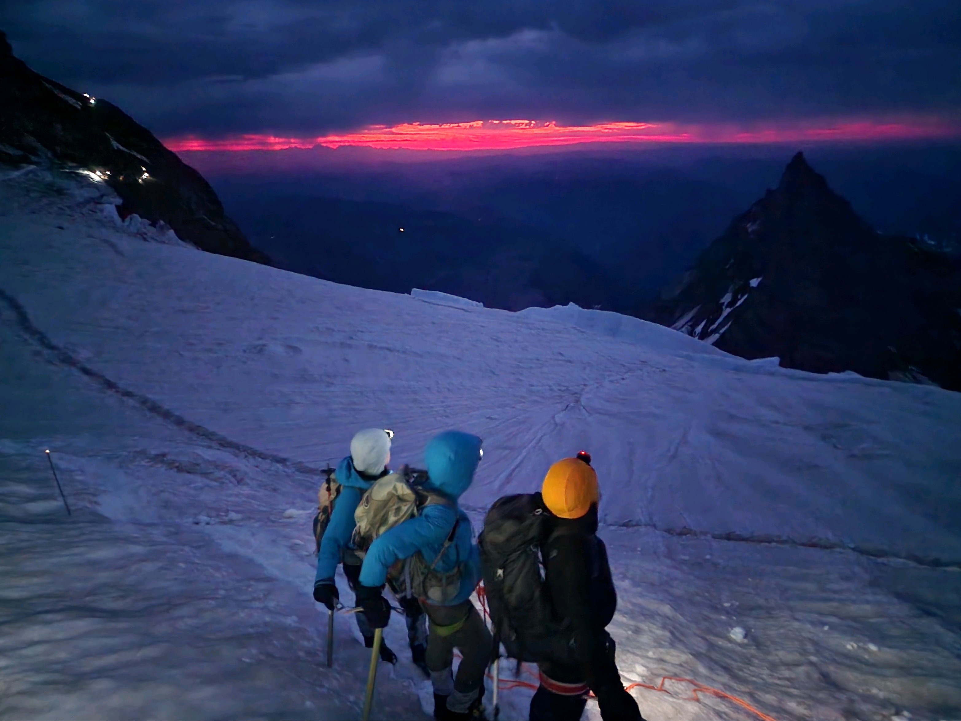 Three climbers on a snowy mountain slope at dawn with headlamps and backpacks, watching a pink sunrise.