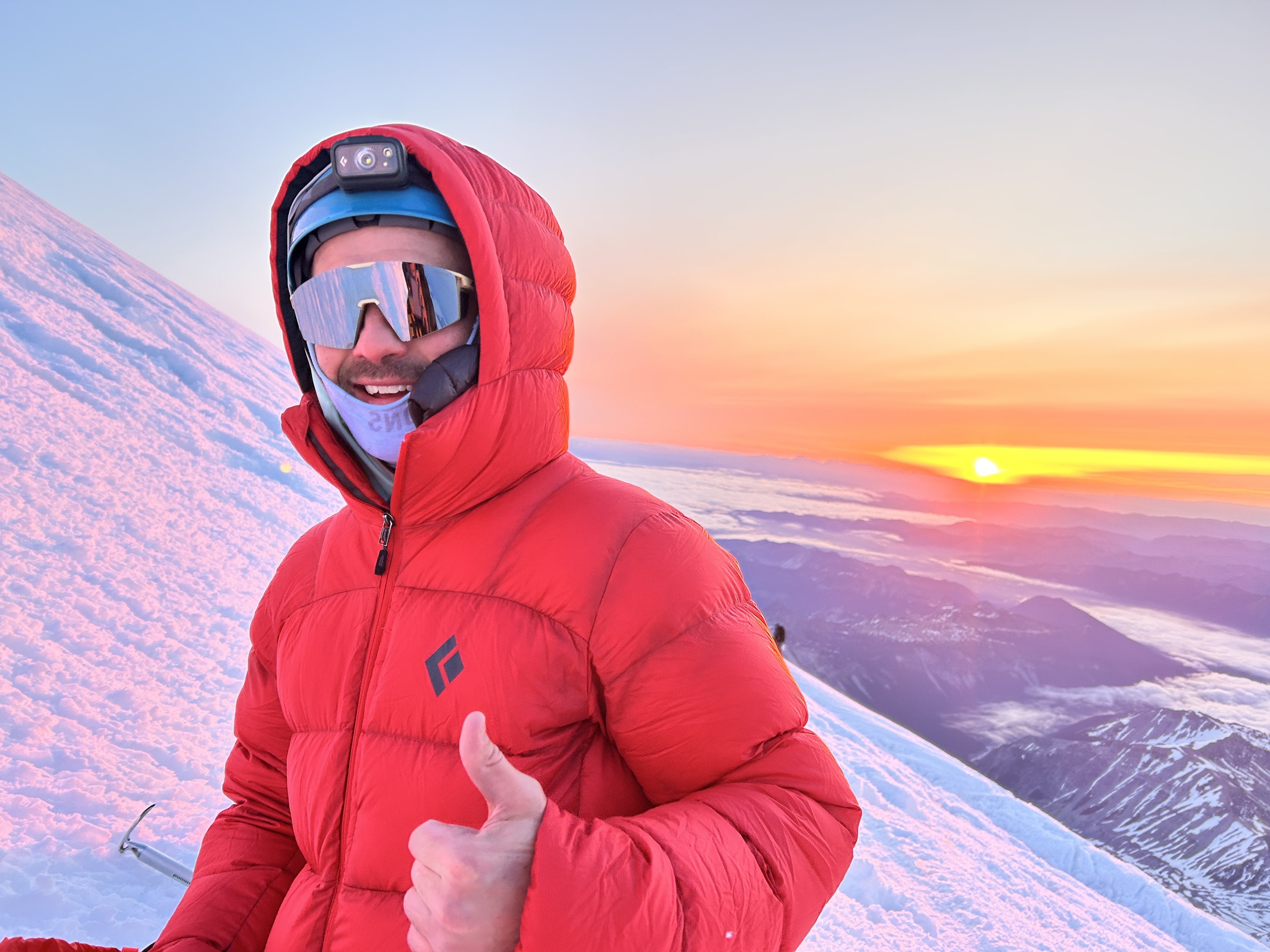 Person in red jacket and helmet with headlamp giving thumbs up on snowy mountain at sunrise.