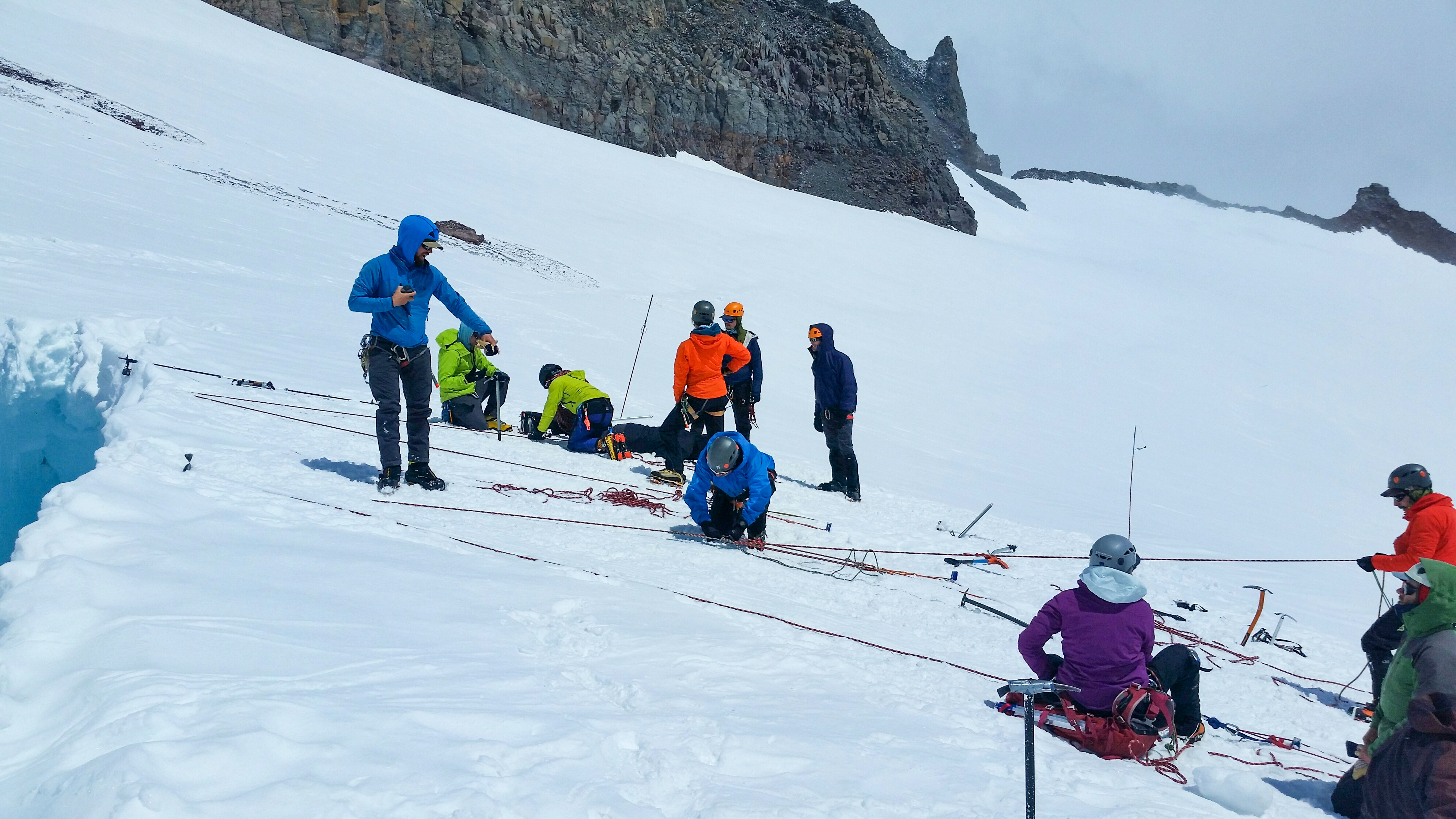 Group of climbers in helmets and jackets training rope rescue on snowy mountain slope near rocky cliff.