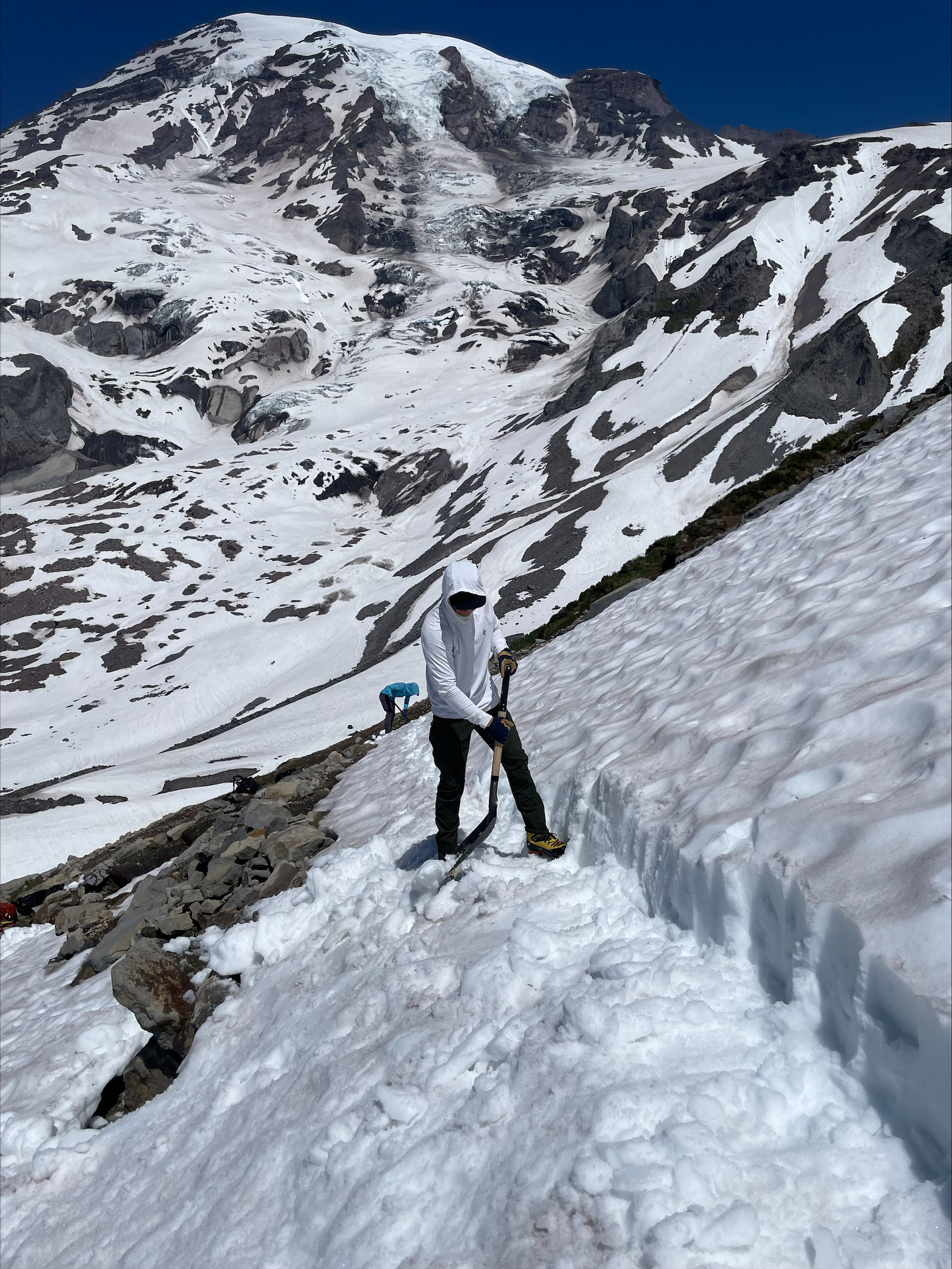 Person in white hoodie using a shovel on a snowy mountain slope with rocky patches and clear blue sky.