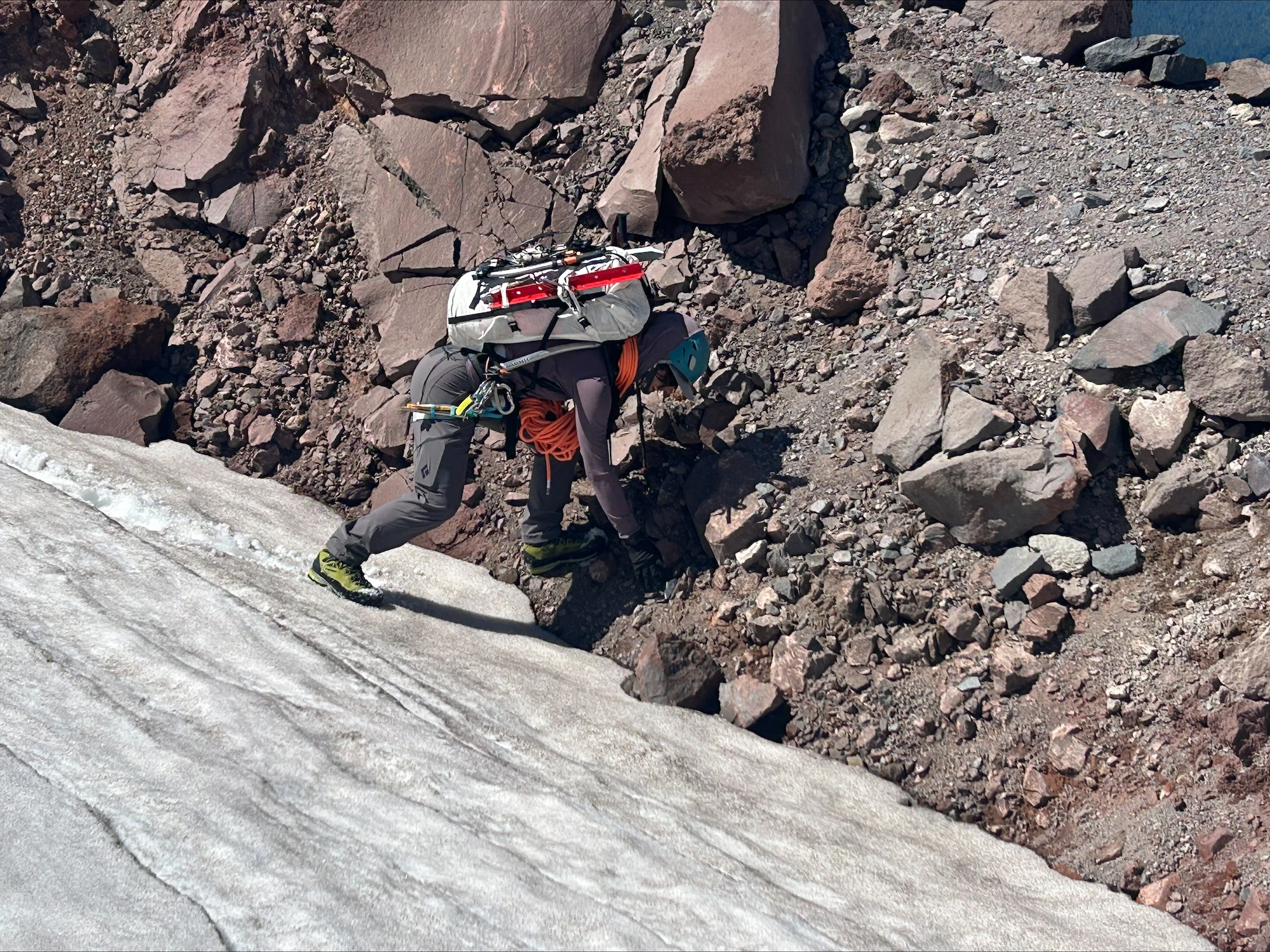 Climber wearing helmet and gear ascending a rocky, snowy mountain slope with a rope coiled around the body.