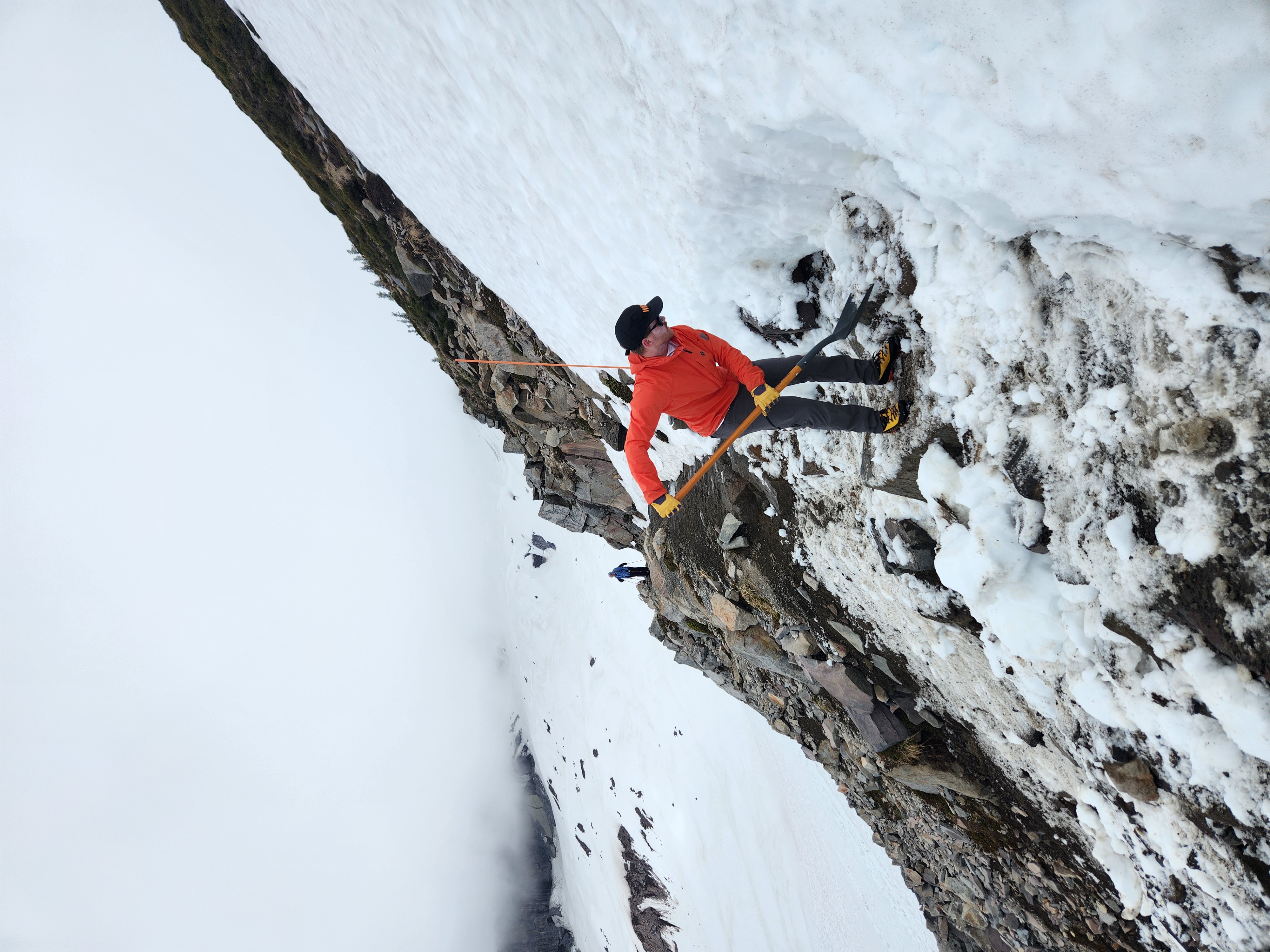 Person in orange jacket shoveling snow on a rocky mountain slope with another hiker in the background.
