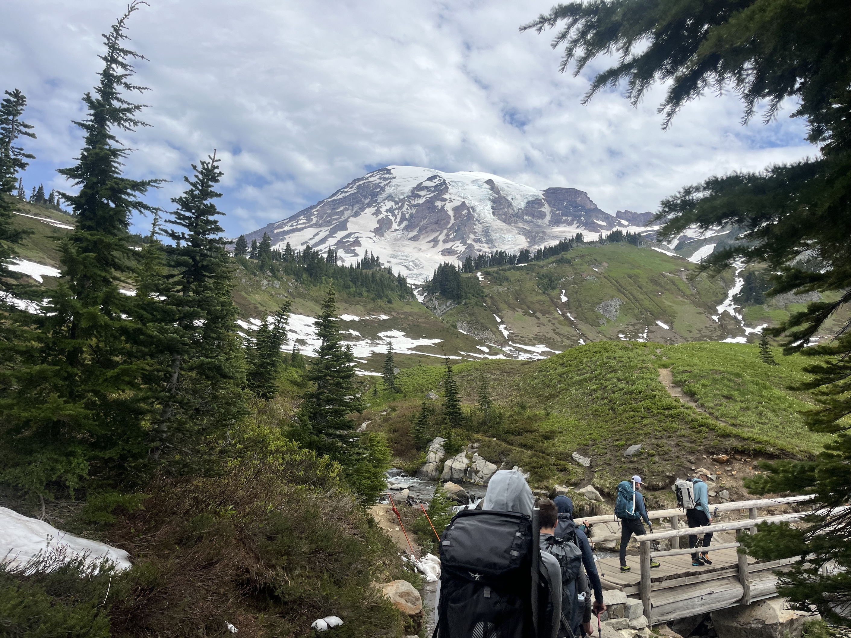 Hikers with backpacks crossing a wooden bridge with snow patches and a snow-capped mountain in background.