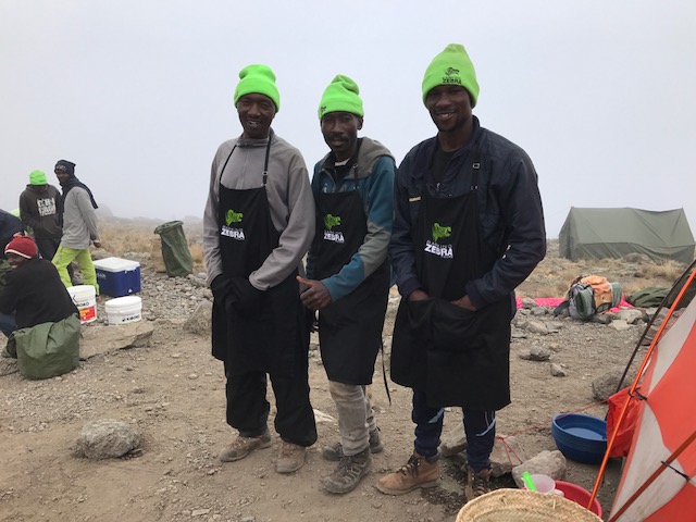 Three men in neon green hats and black aprons standing on rocky ground with tents in the foggy background.