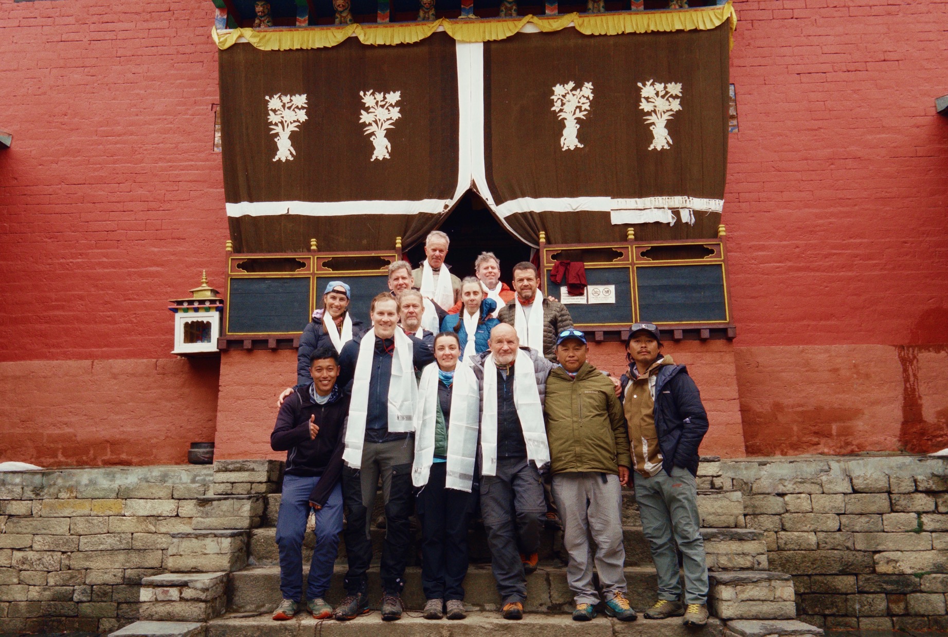 Group of hikers wearing white scarves posing on stone steps in front of red temple with decorated brown curtains.