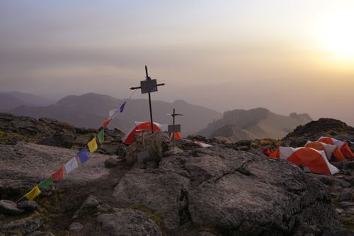 Mountain campsite with orange tents near a memorial cross at sunset with hazy hills in the background.