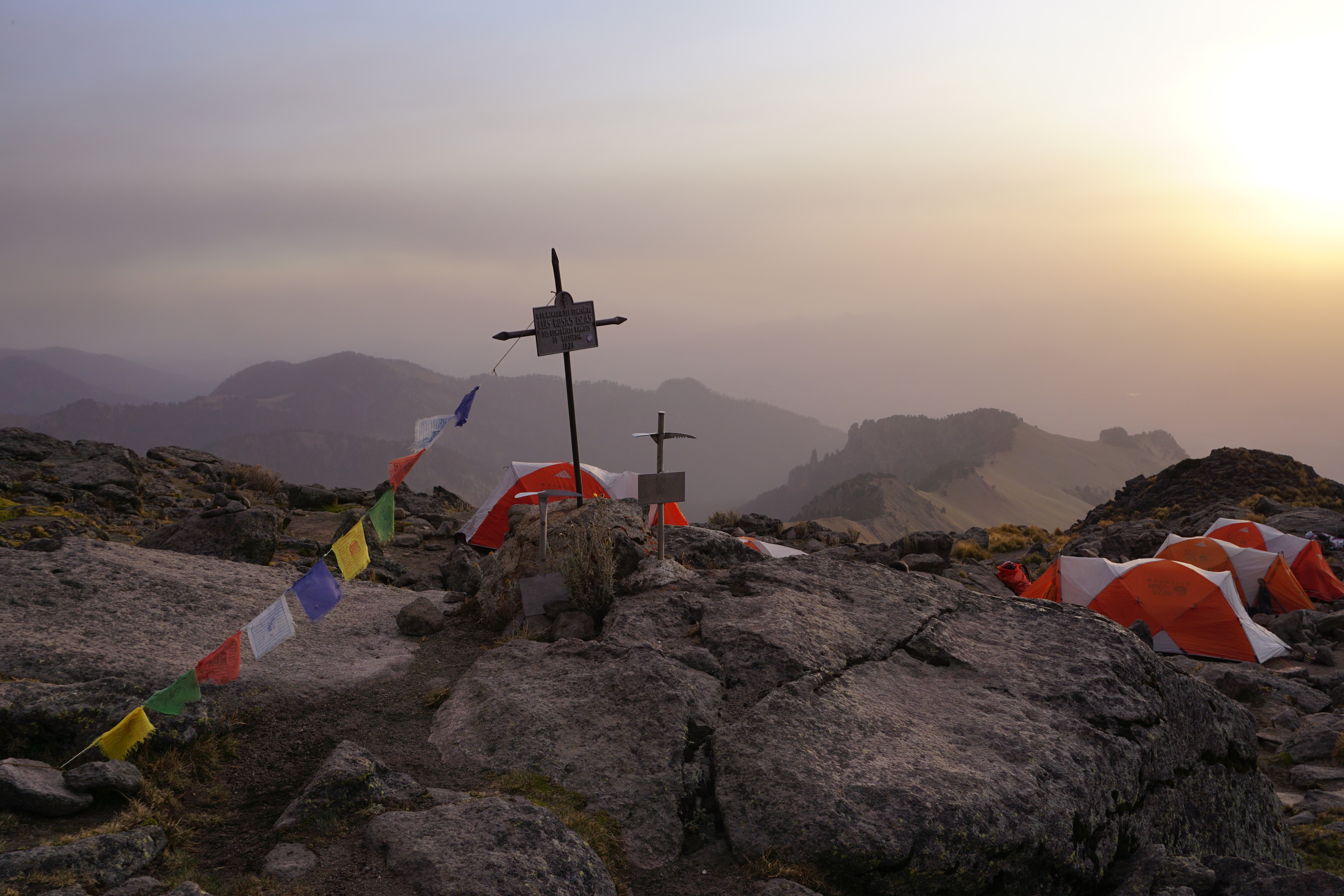 Mountain campsite with orange tents near a memorial cross at sunset with hazy hills in the background.