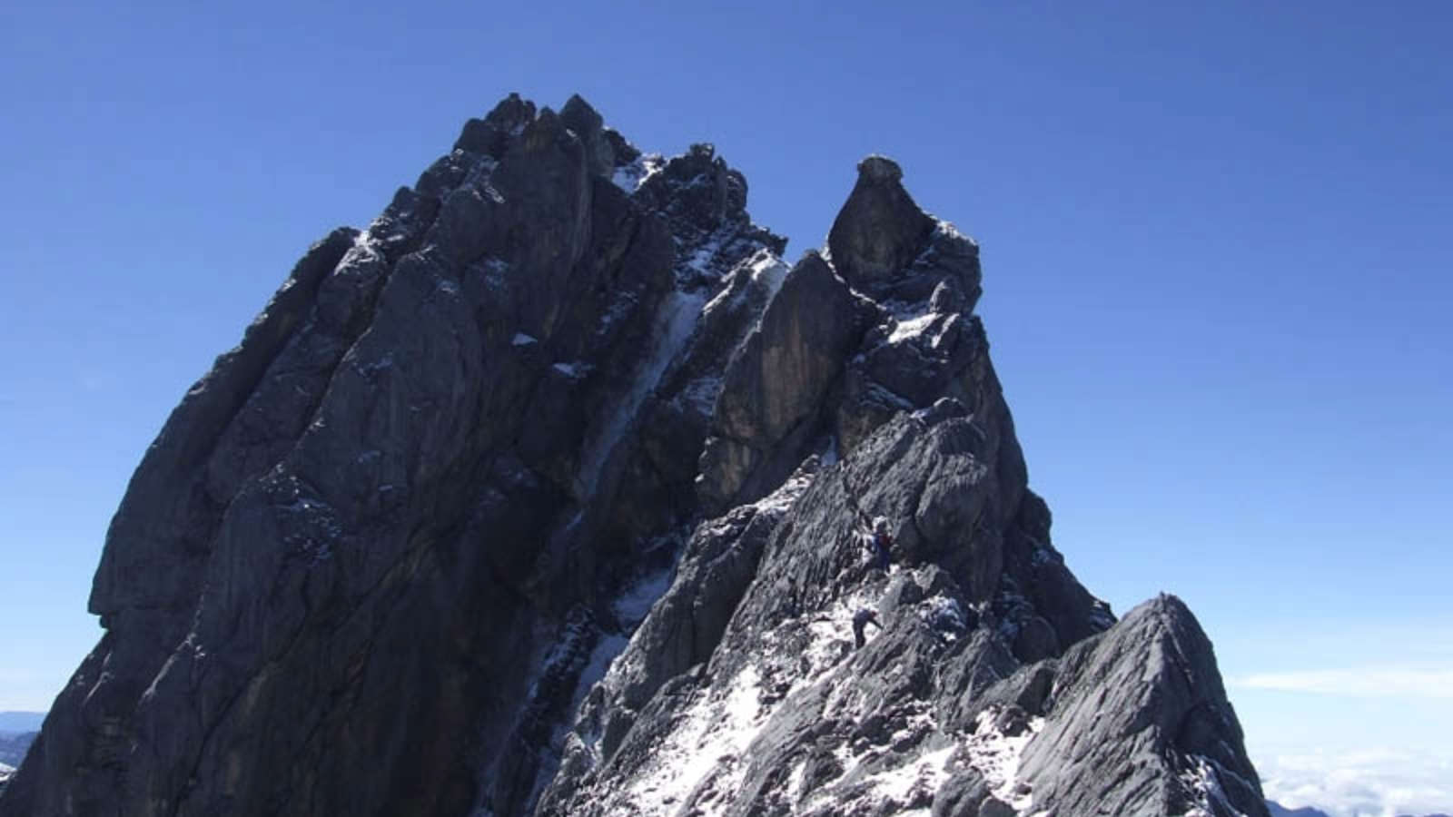 Climbers ascending a steep rocky mountain ridge with patches of snow under a clear blue sky.