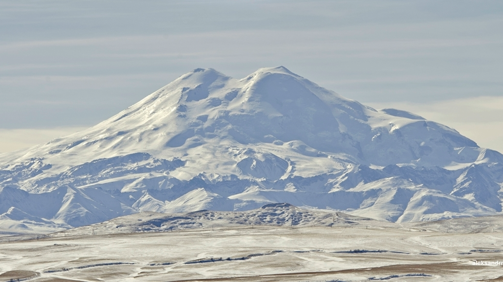 Snow-covered mountain with a broad peak under a cloudy sky and snowy plain in the foreground.