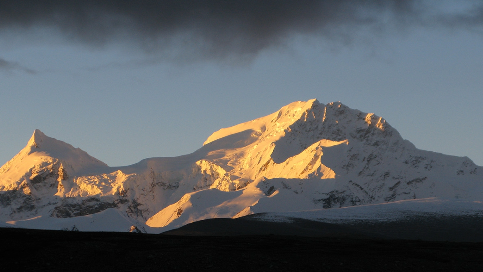 Snow-covered mountain peaks bathed in warm sunlight under a cloudy blue sky.