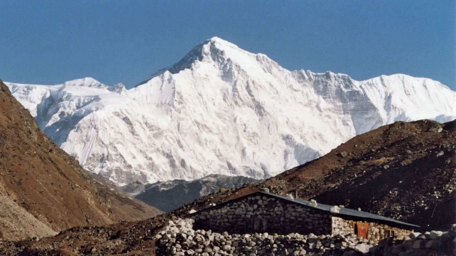Snow-covered mountain peak behind rocky hills with a stone shelter in the foreground.