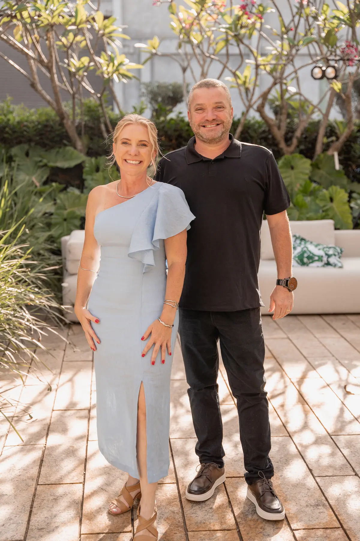 Smiling man and woman pose outdoors on a sunny patio with plants and sofa in the background.